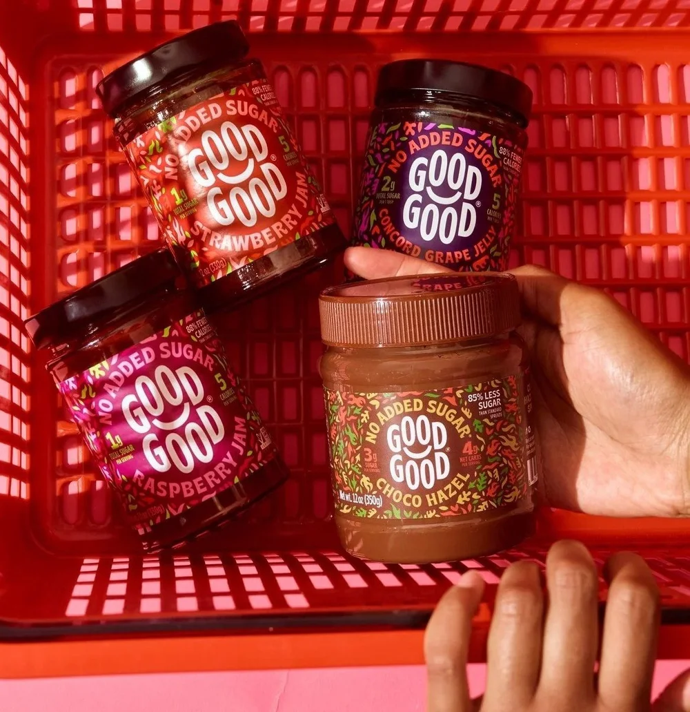 Hands placing Good Good jam jars in a red shopping basket on pink, retail CPG photography