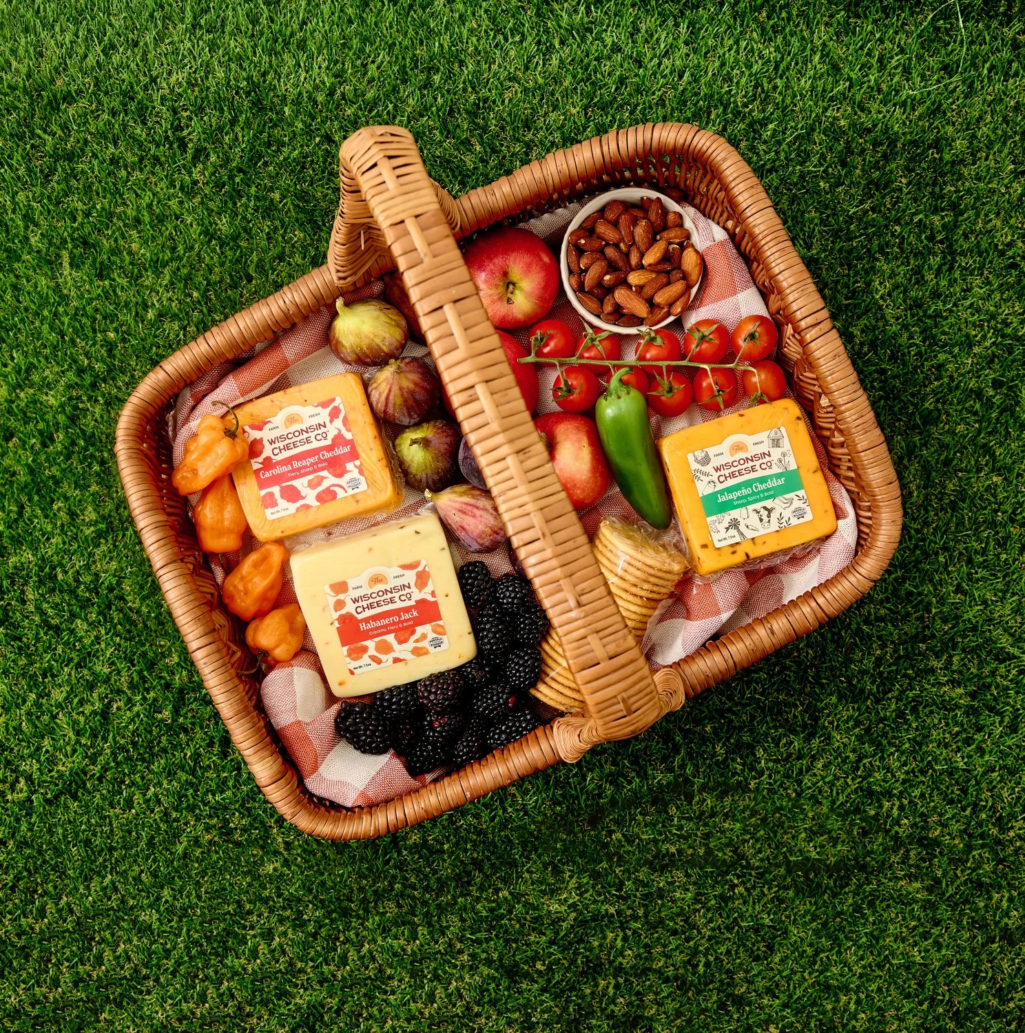 A picnic basket filled with cheese, figs, tomatoes, almonds, blackberries, and pepper peppers, placed on green grass.