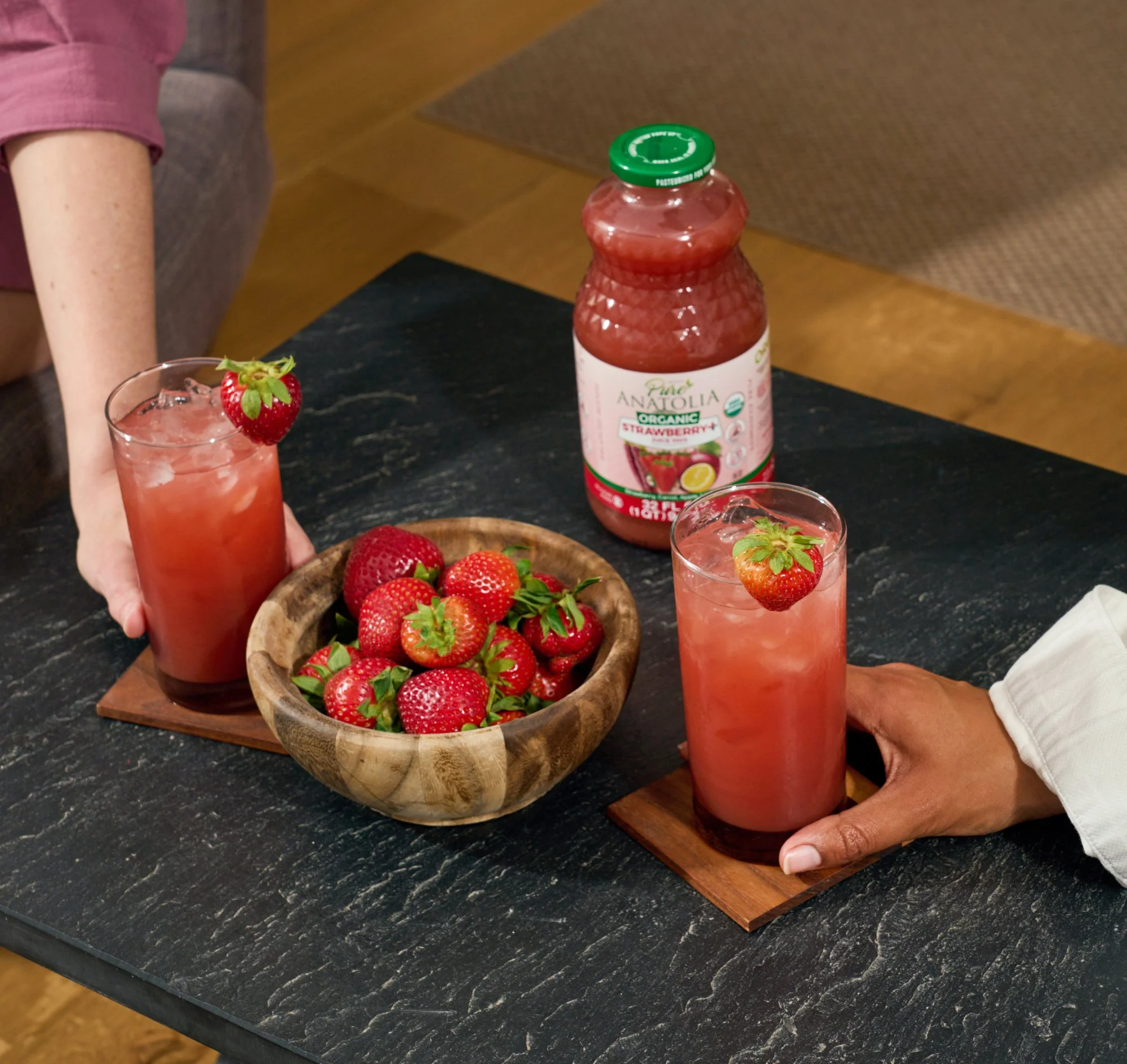 A person wearing a white shirt is holding a tall glass of strawberry juice garnished with a strawberry on the rim. On the black countertop, there is a wooden bowl filled with fresh strawberries and a bottle of organic strawberry juice with a green ca