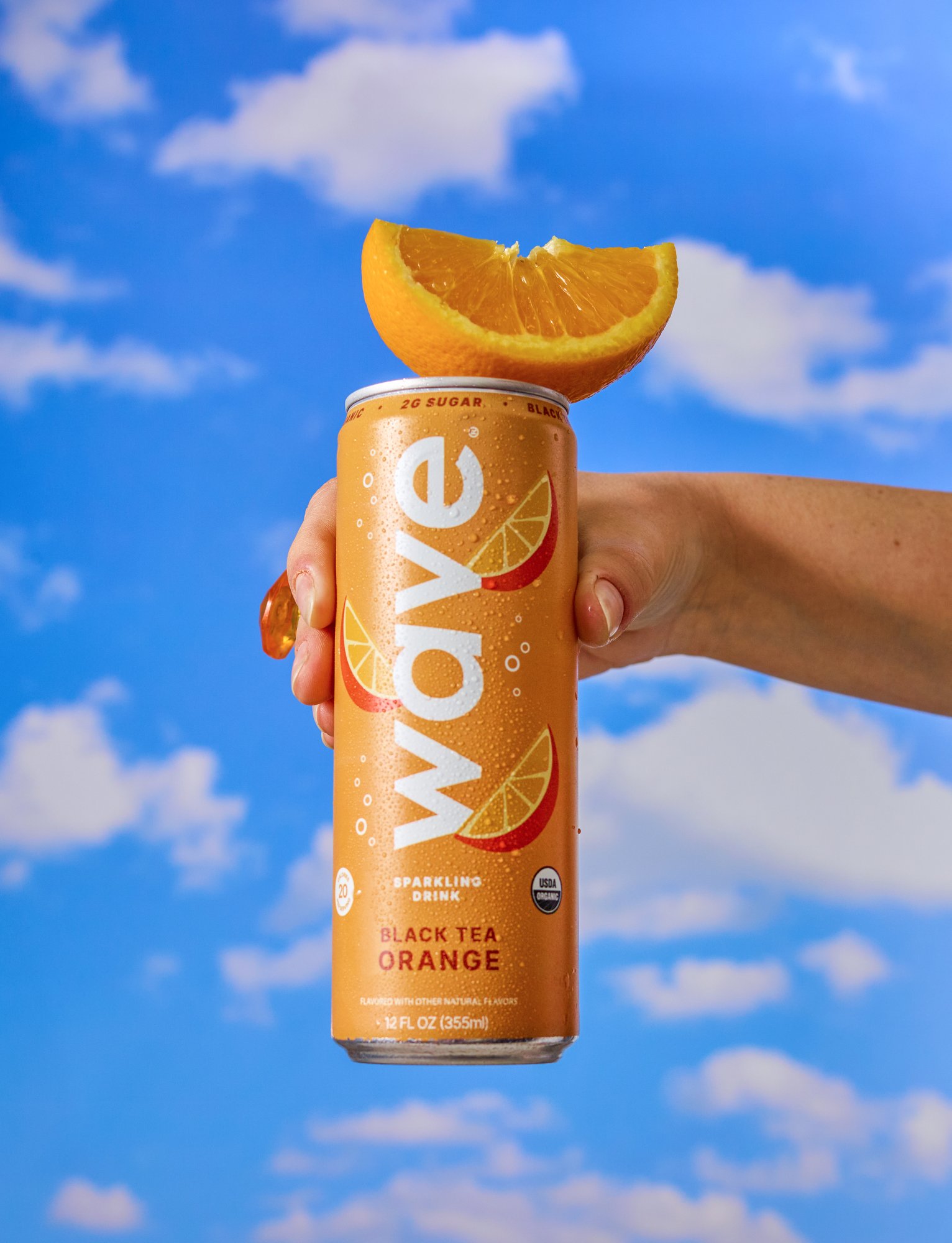 A hand holding a can of Sparkling Orange Black Tea beverage with a slice of orange on top against a blue sky with clouds.