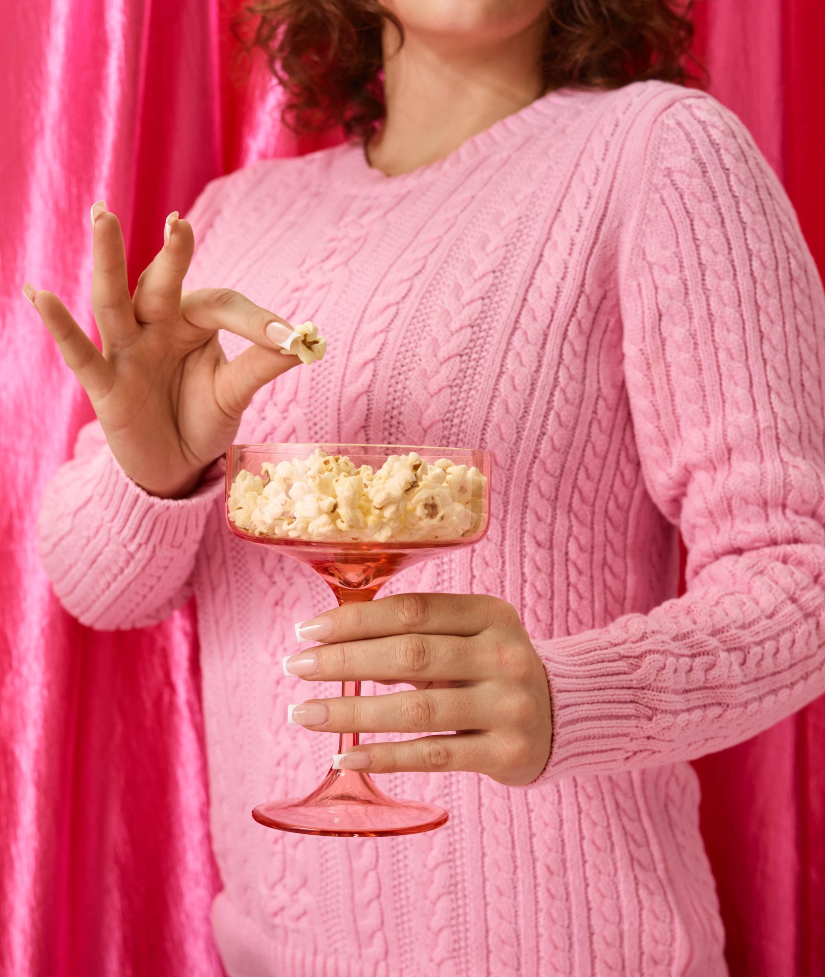 A woman in a pink cable-knit sweater holding a glass of popcorn with a pink background.