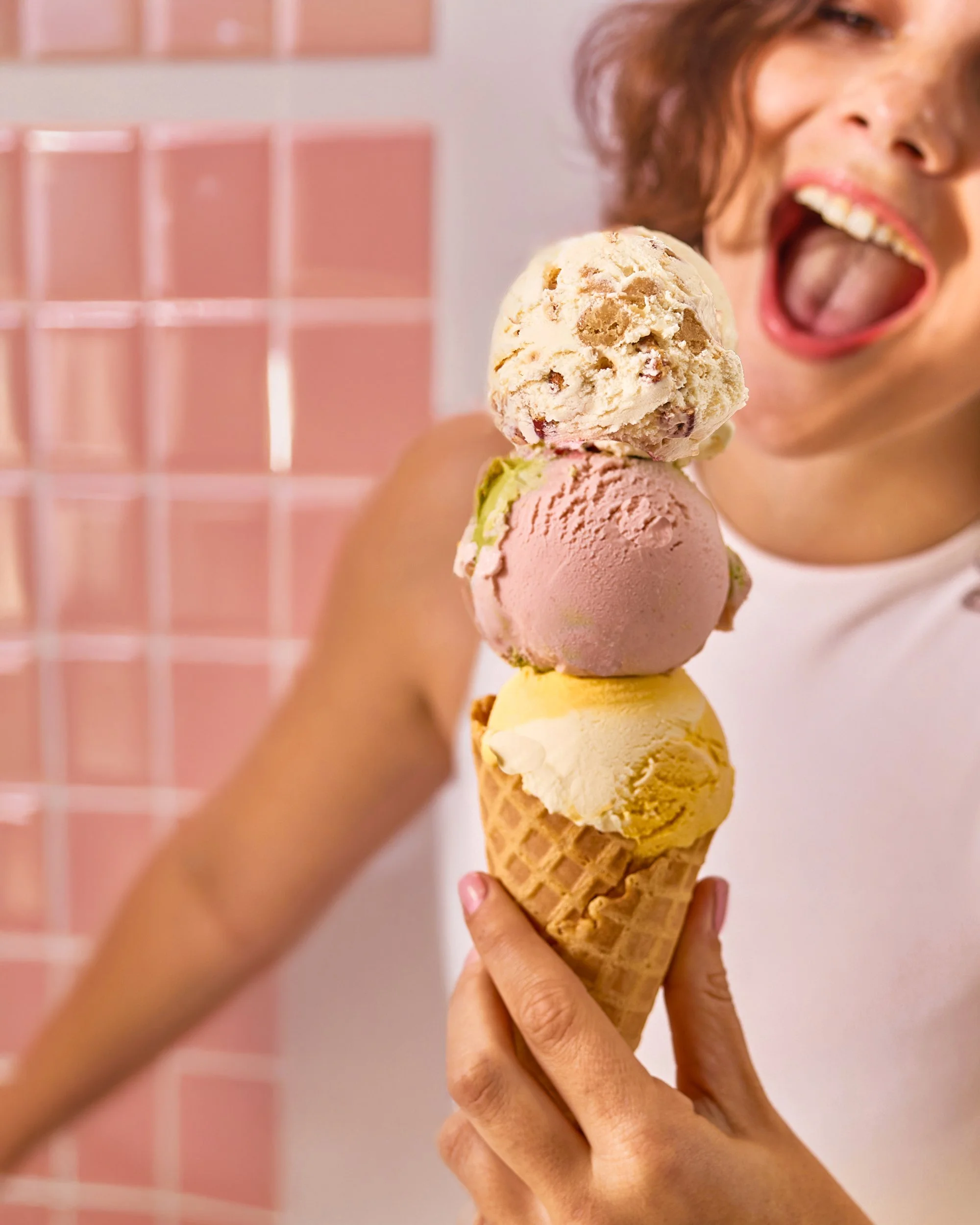 A person holding a waffle cone with three scoops of ice cream in front of a pink tile wall, opening their mouth and reaching towards the ice cream.
