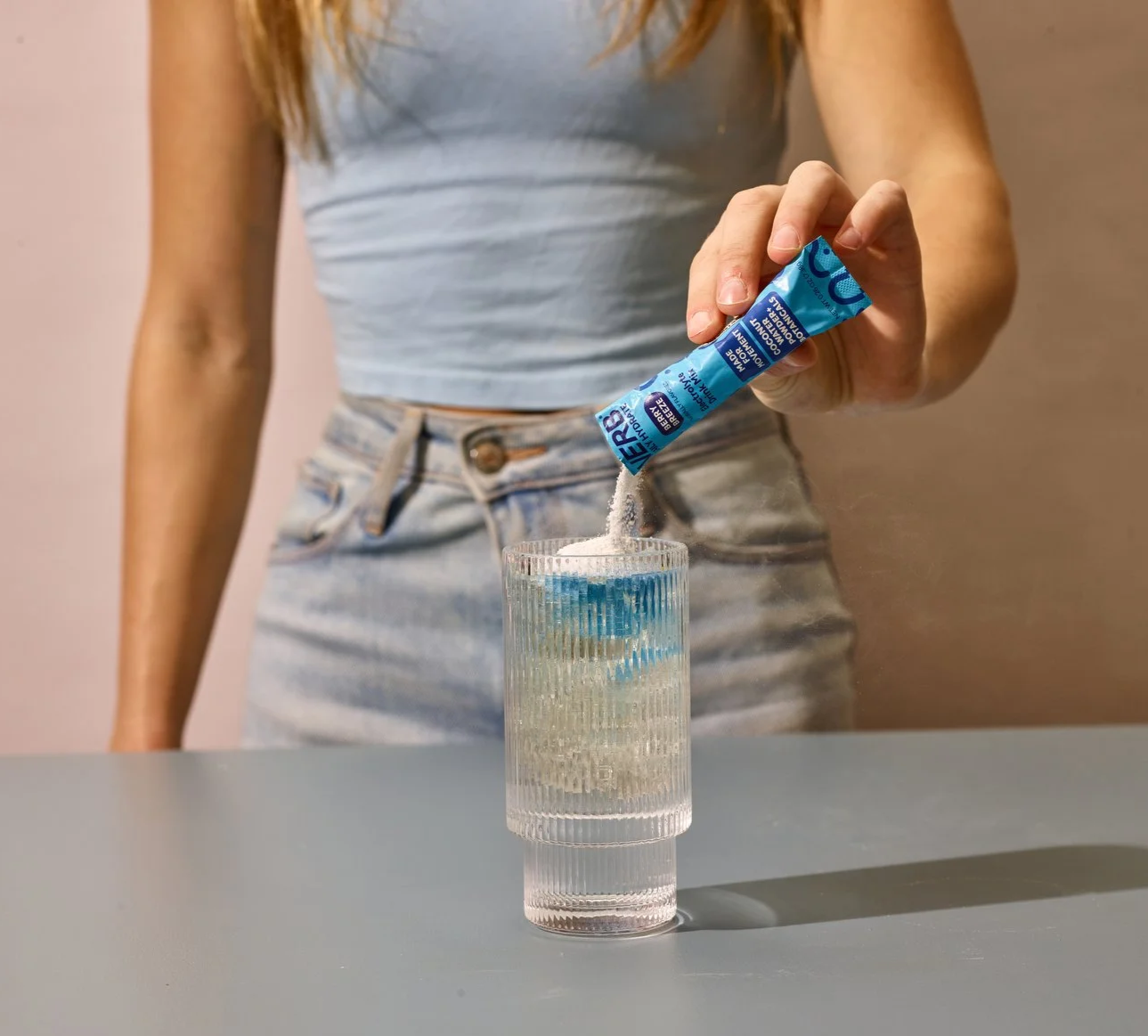 A woman in a light blue sleeveless top and high-waisted jeans is pouring powder from a small blue packet into a clear glass of water on a gray table.