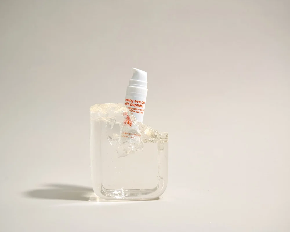 A white skincare tube labeled 'Soothing eye gel with peptides' sitting on a block of ice inside a glass container on a plain background.