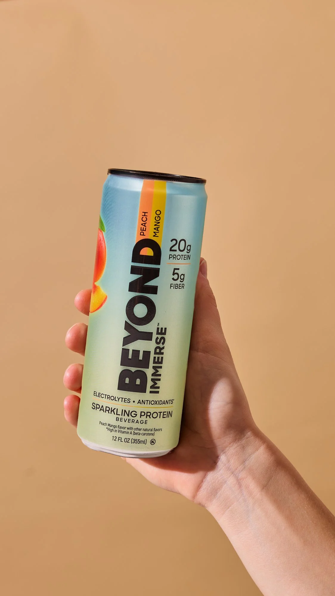 A person holding a light blue can of BEYOND IMMERSION sparkling protein beverage with peach mango flavor against a beige background.