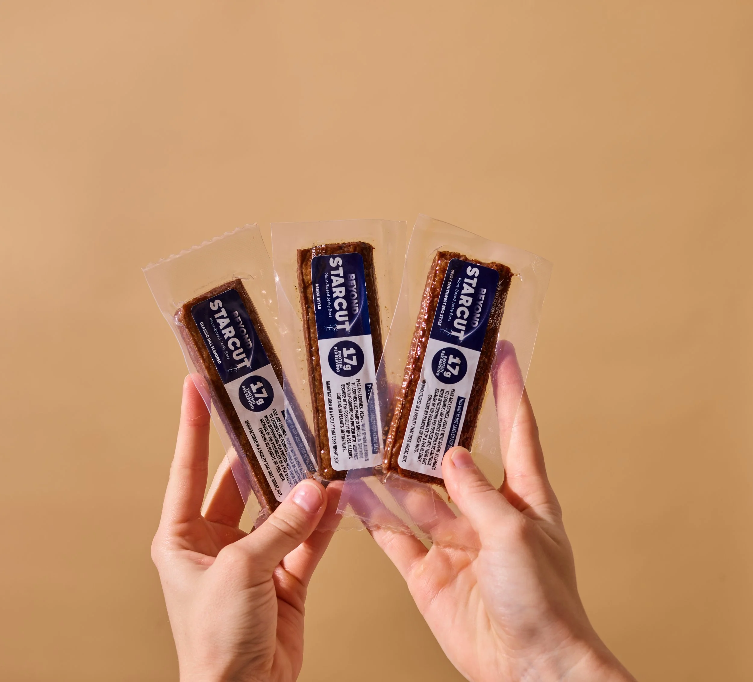 Person holding three packaged beef jerky sticks against a plain beige background.