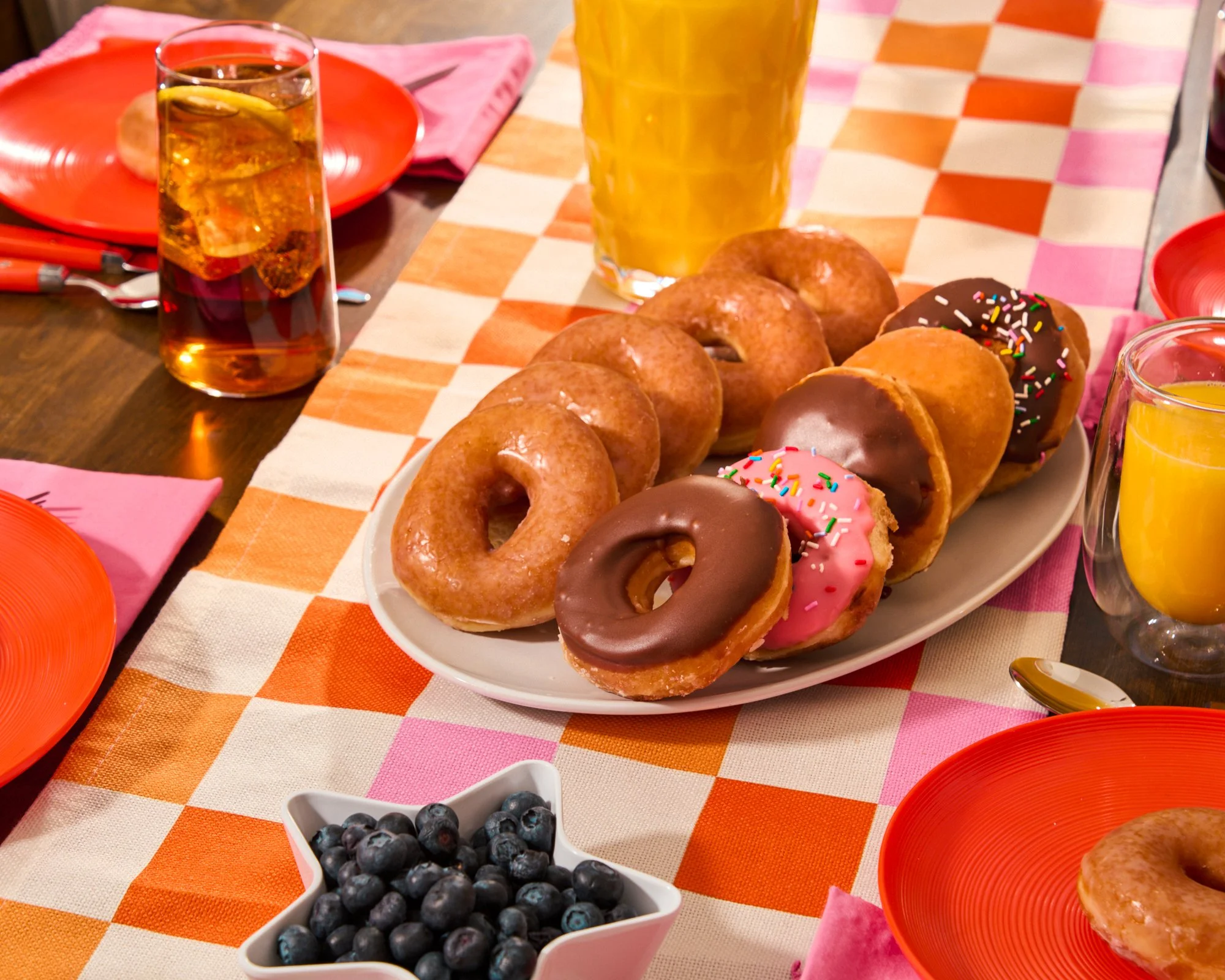 Assorted donuts with pink and chocolate icing and sprinkles on a white platter, blueberries in a star-shaped dish, glasses of orange juice, glasses of iced tea, and orange and pink plates on a checkered orange and white tablecloth.