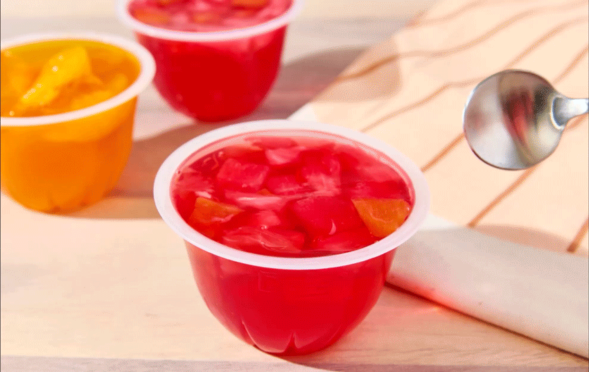 Side view of a red fruit jelly cup with fruit chunks, with other fruit jelly cups in the background on a light wood surface.