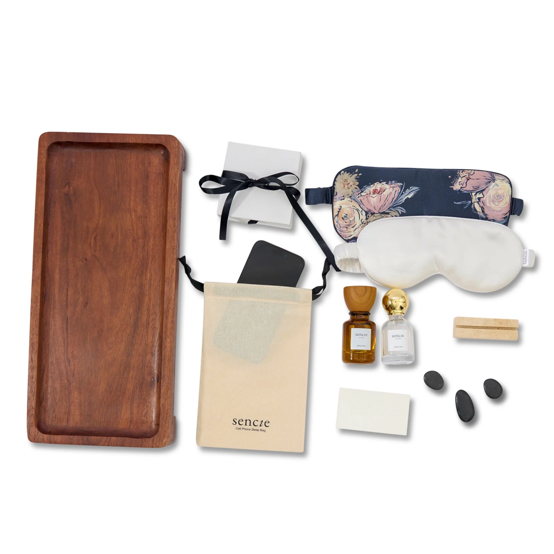 Collection of sleep accessories including a wooden tray, sleep mask, herbal eye pillow, cell phone sleep bag, small white box, two perfume bottles, an eraser, two black stones, and a sheet of paper.