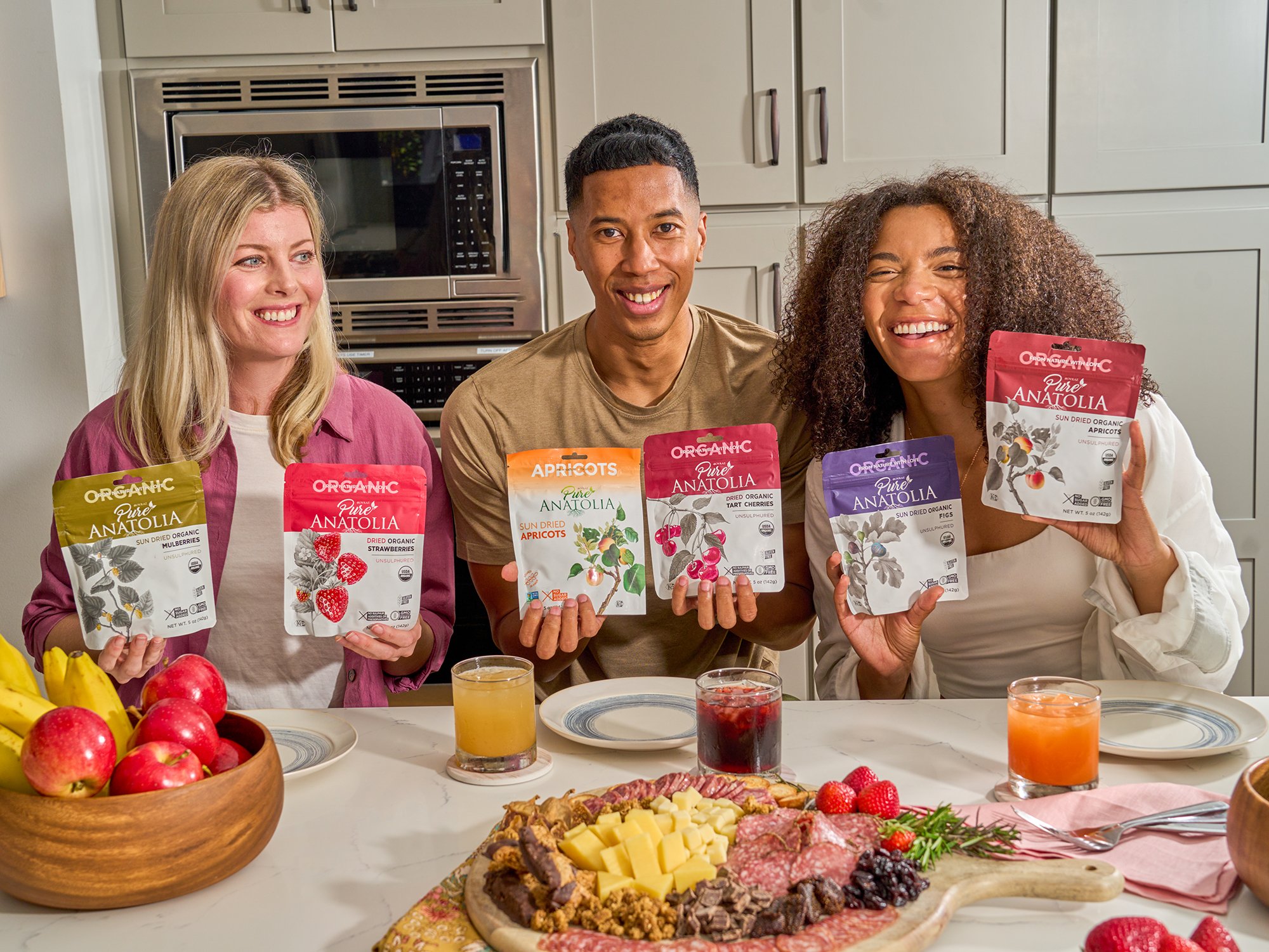 Three people standing behind a table with a variety of food, including apples, cheese, meats, and finger foods, in a kitchen setting, smiling and holding packages of dried organic fruit.