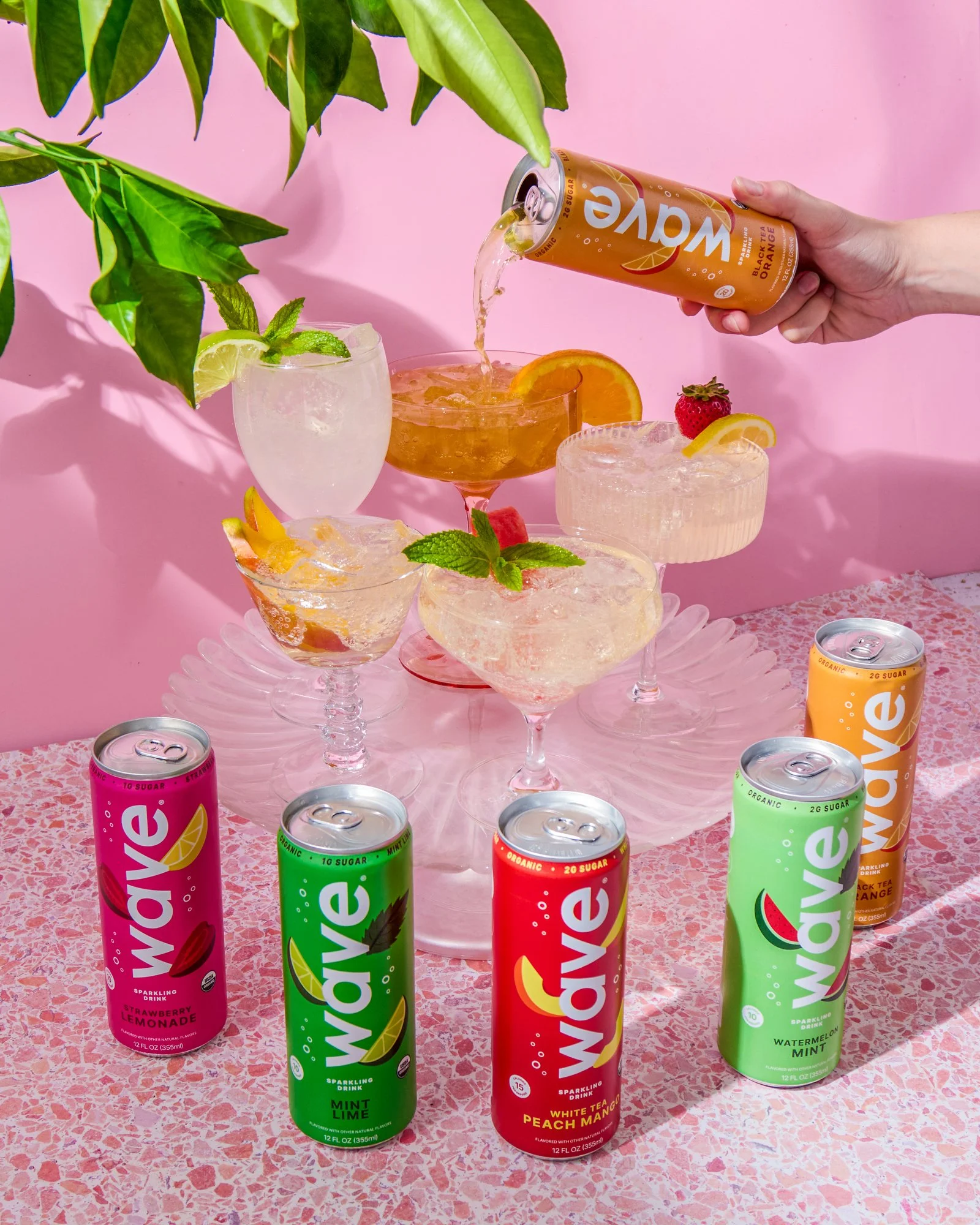 A pink textured table with cans of Wave sparkling drink in strawberry lemonade, mint lime, white tea peach mango, and watermelon mint flavors. A clear glass with lime and mint is on the left, and a crystal dessert dish with orange slice and mint is n