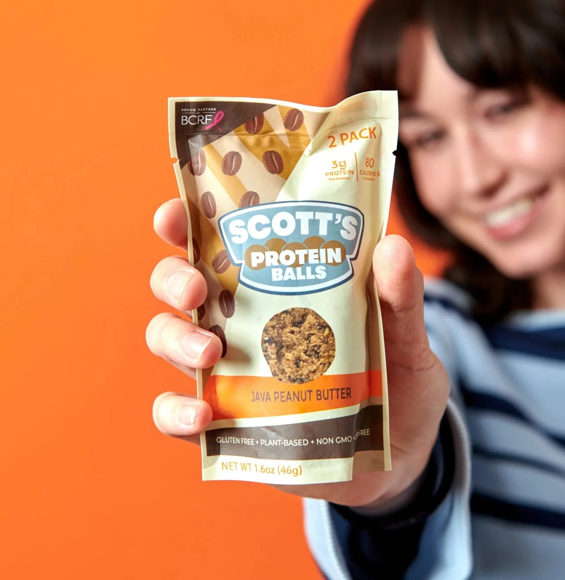 Hand holding a Scott's protein balls bag toward camera on orange, CPG lifestyle photography