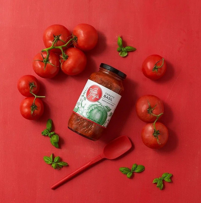 Pasta sauce jar surrounded by tomatoes and basil on red, CPG food photography