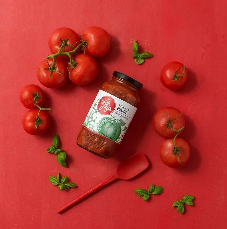 Pasta sauce jar surrounded by tomatoes and basil on red, CPG food photography