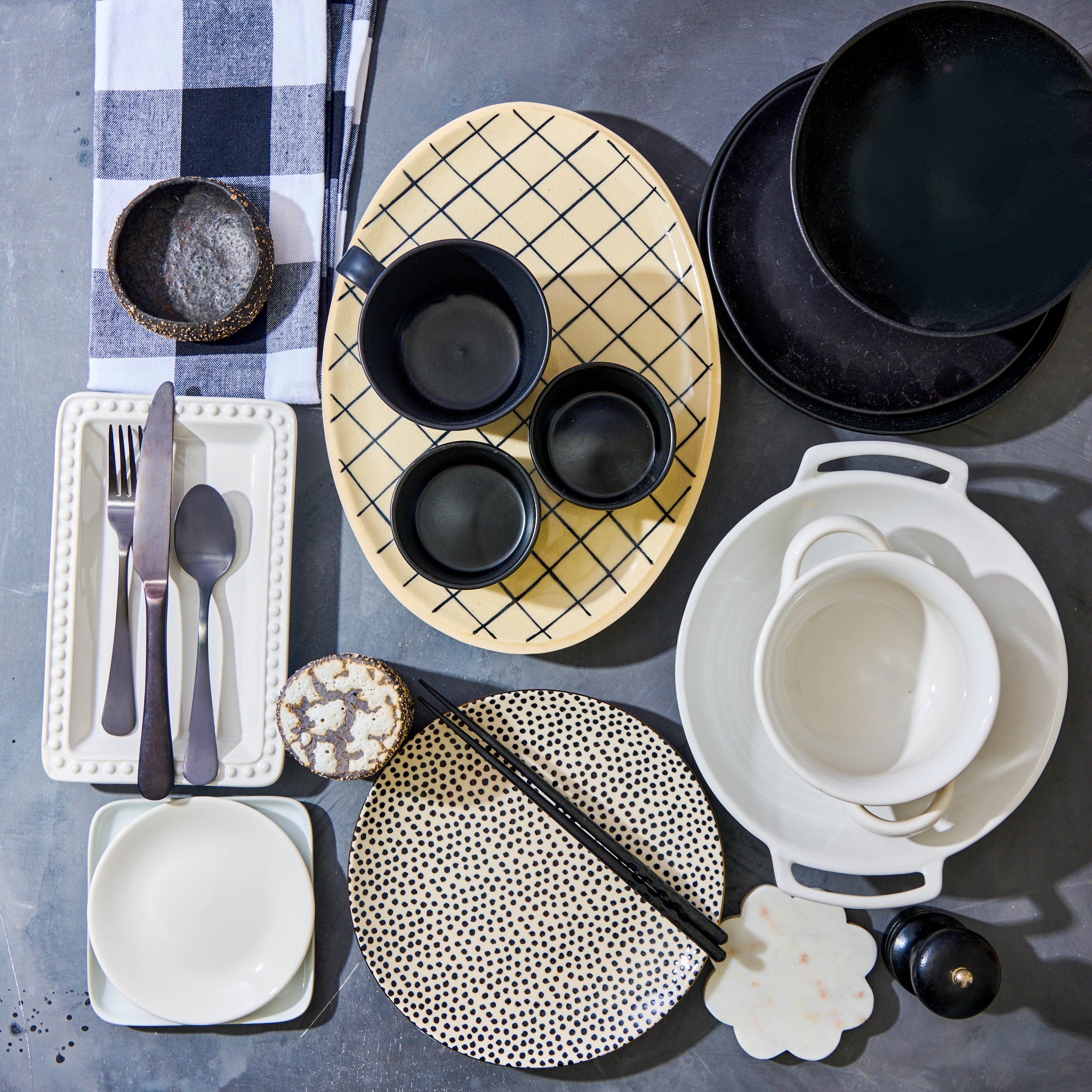 Various black, white, and patterned dinnerware including plates, bowls, cups, and utensils arranged on a table with a checkered napkin.