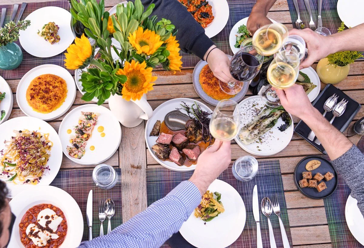 Overhead of friends toasting wine over shared restaurant dishes and sunflowers