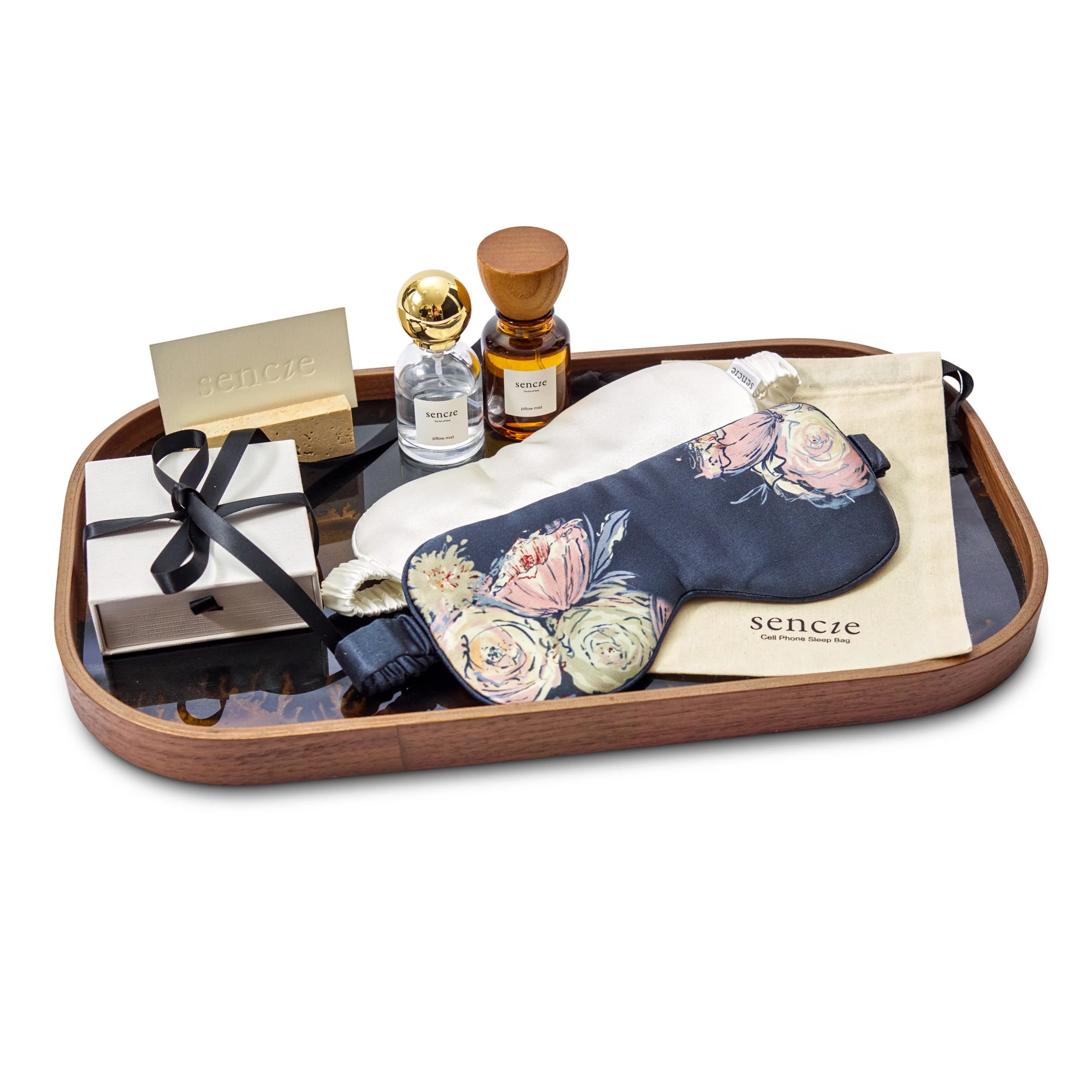 A wooden tray holding sleep accessories including a floral eye mask, a cloth bag, a small box with a black ribbon, a soap bar, two bottles of scented oil, and a small pillow.