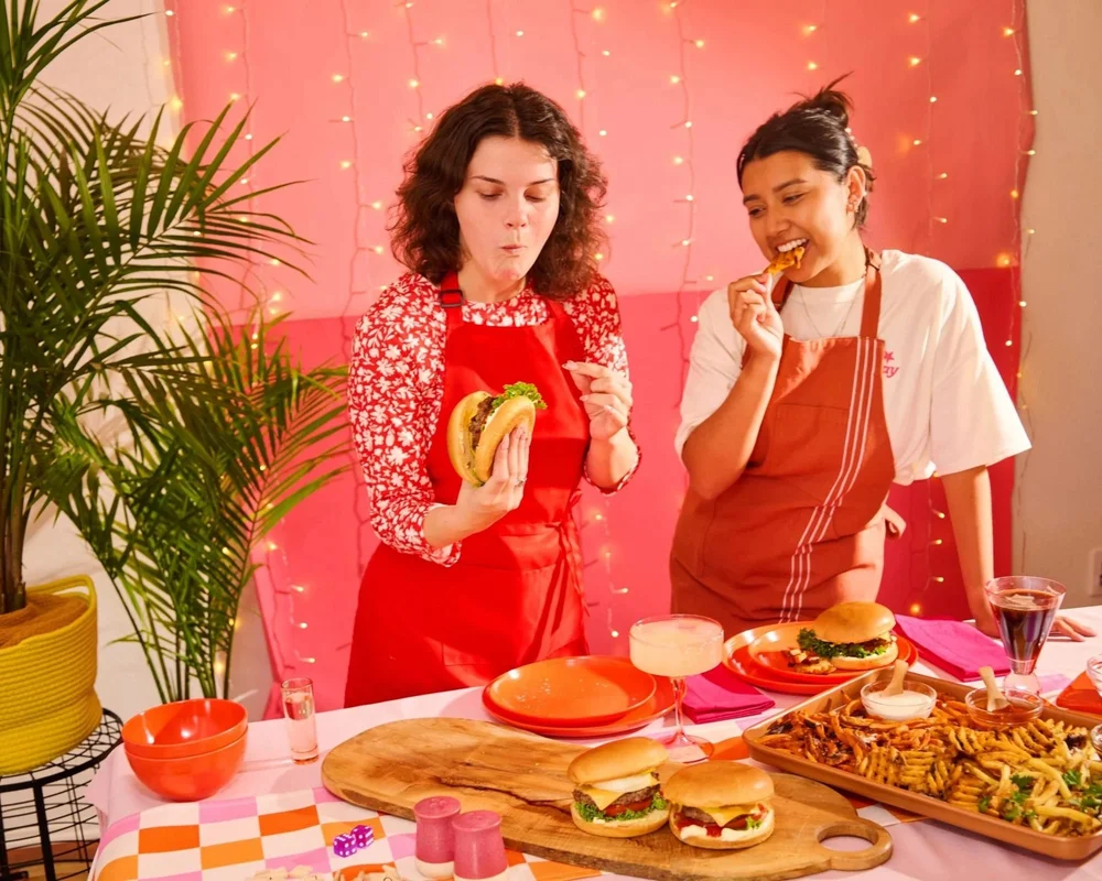Two women tasting burgers at a pink party table, food lifestyle photography