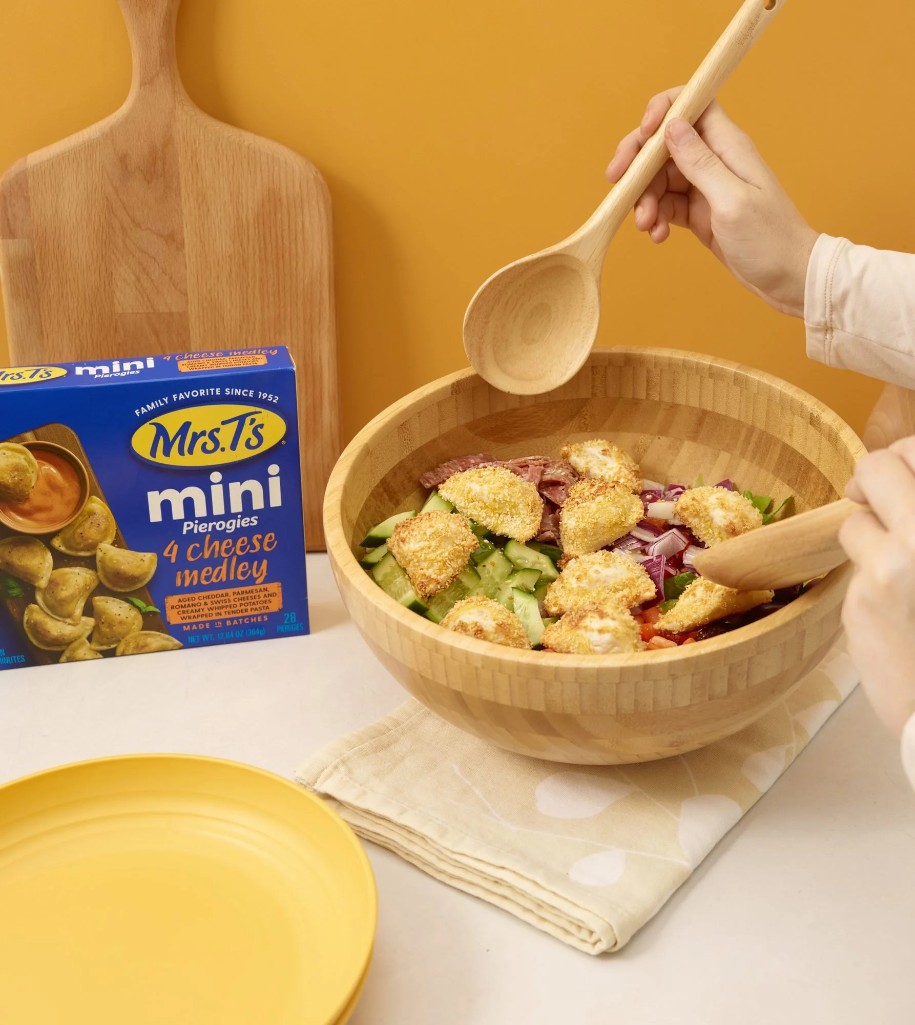 Mrs. T's mini pierogies box with hands tossing a salad on yellow, CPG frozen food photography