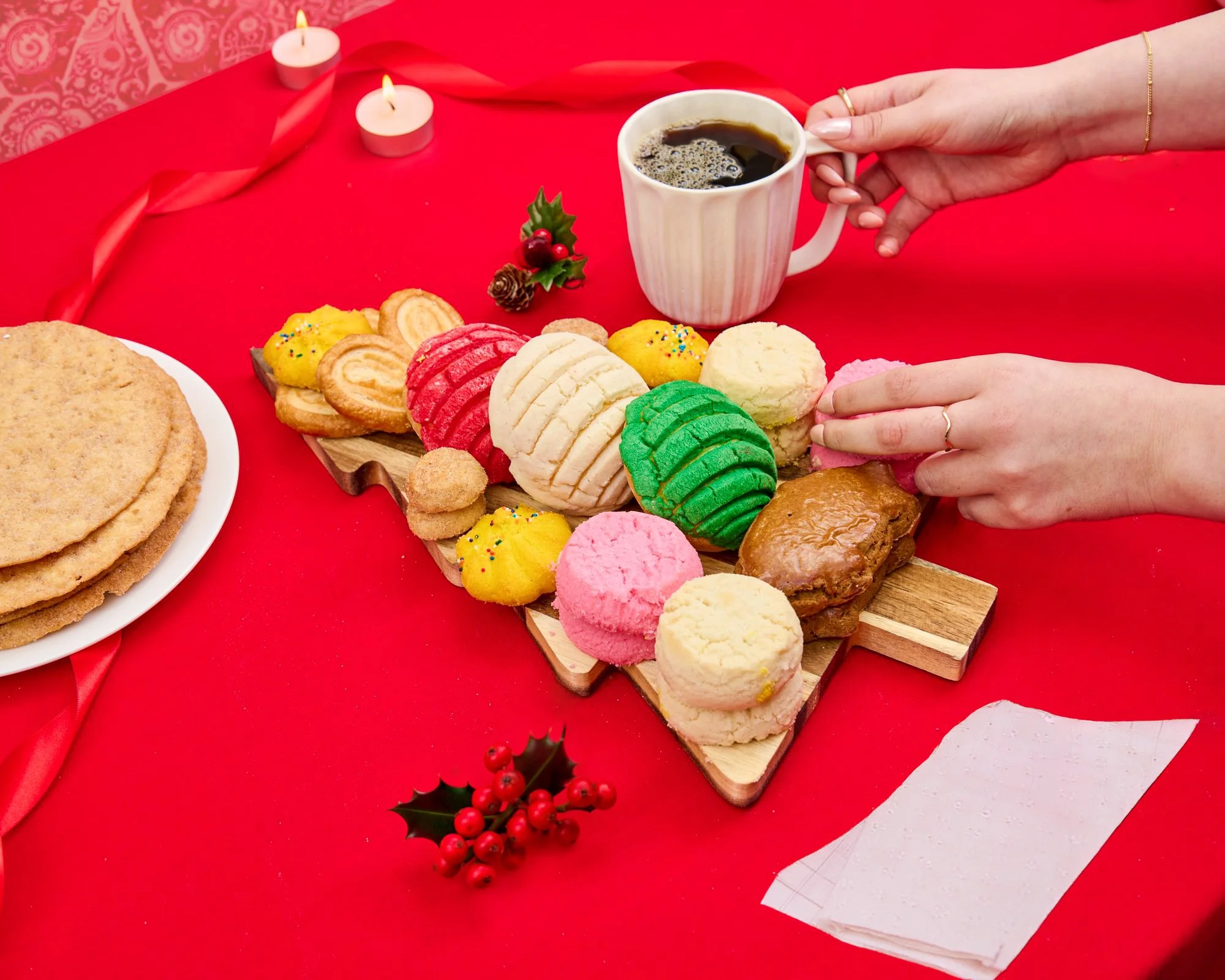Hands reaching for holiday conchas, cookies and coffee on a red table. Seasonal food photography for grocery and bakery campaigns.