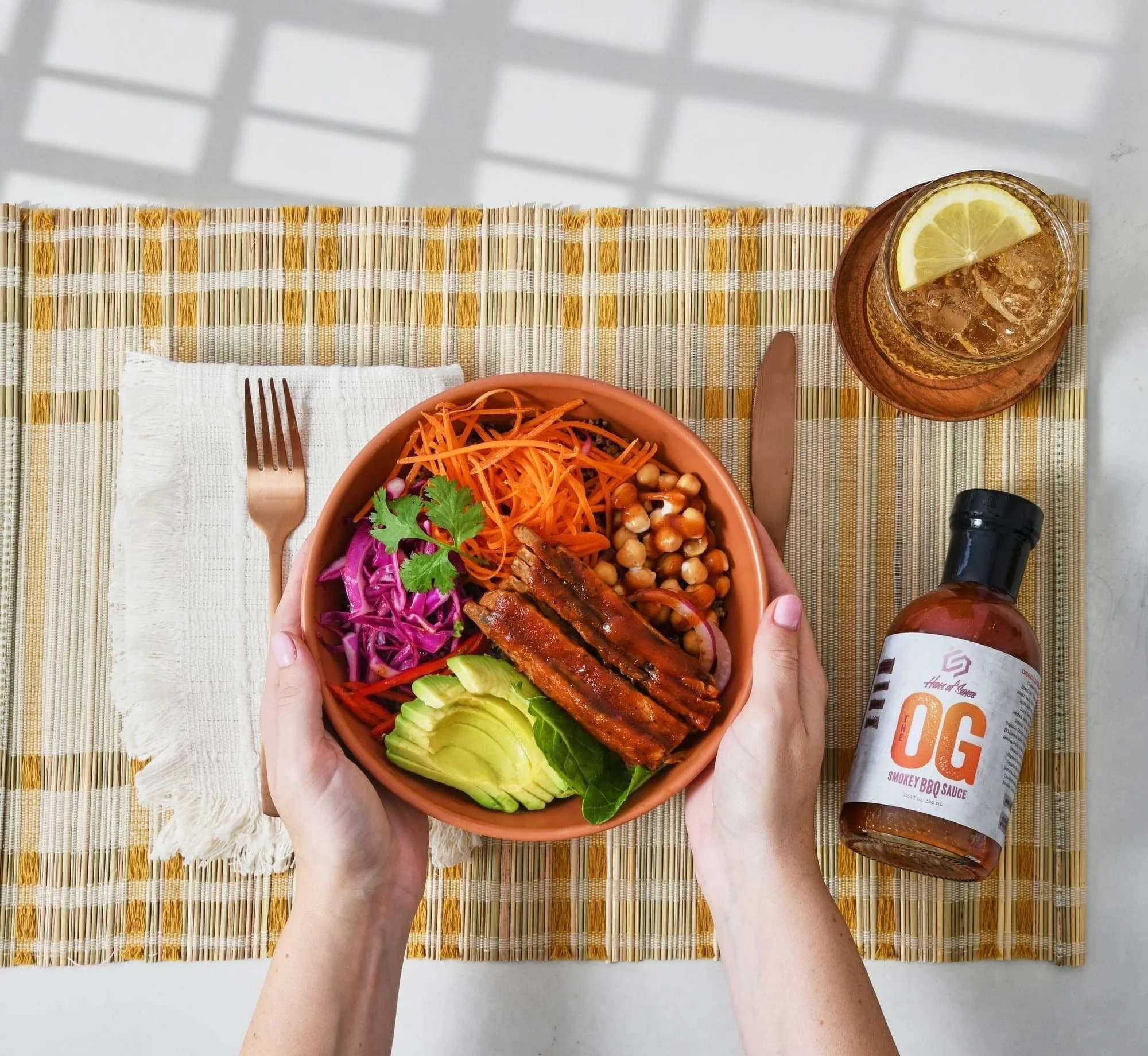 Hands holding a grain bowl with an OG sauce bottle on a plaid cloth. Lifestyle CPG sauce photography.
