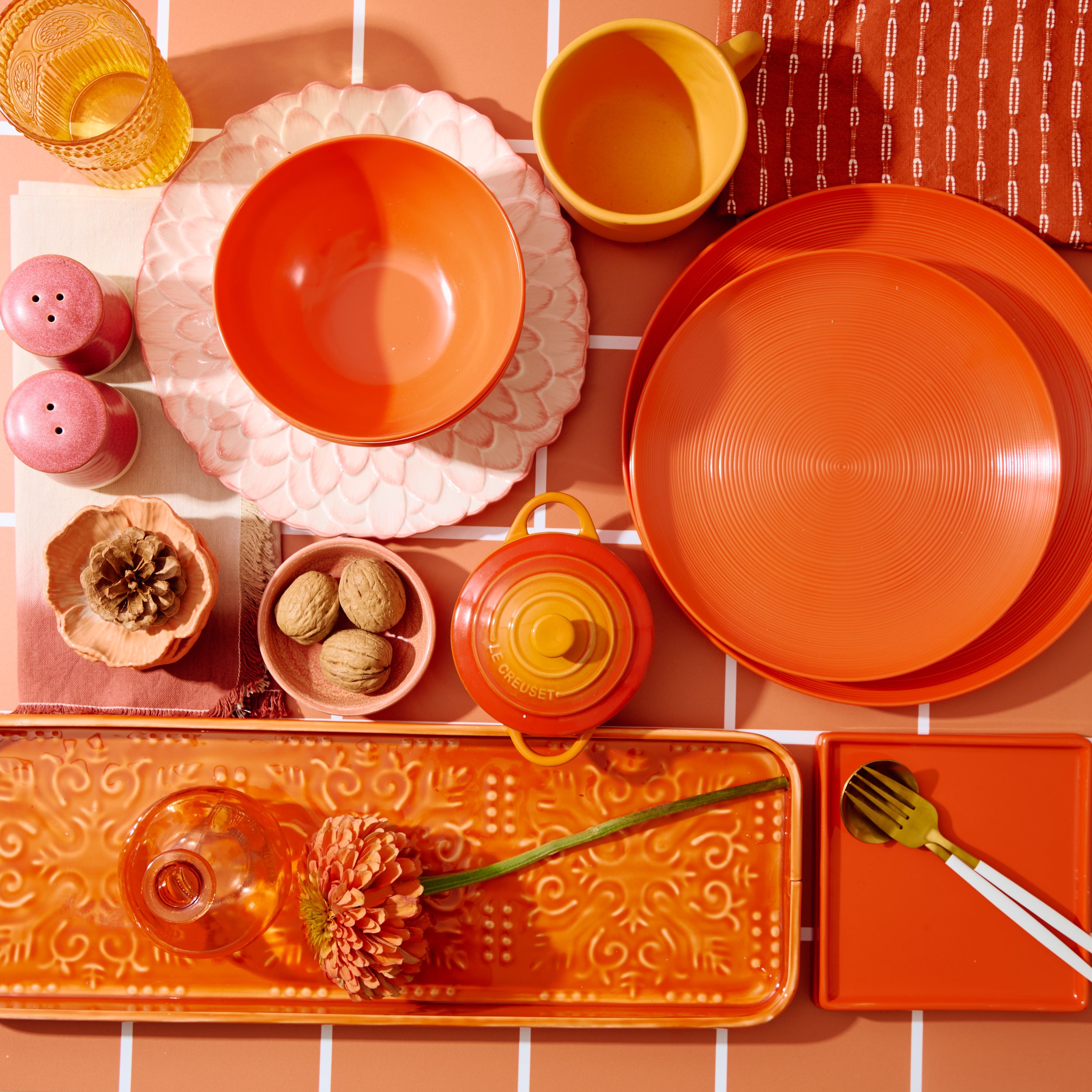 Table set with orange and pink dishes, including bowls, plates, cups, salt and pepper shakers, a small jar with a flower, walnuts, a small pot, a decorative tray with a flower and a glass, and a fork and spoon on an orange tray.