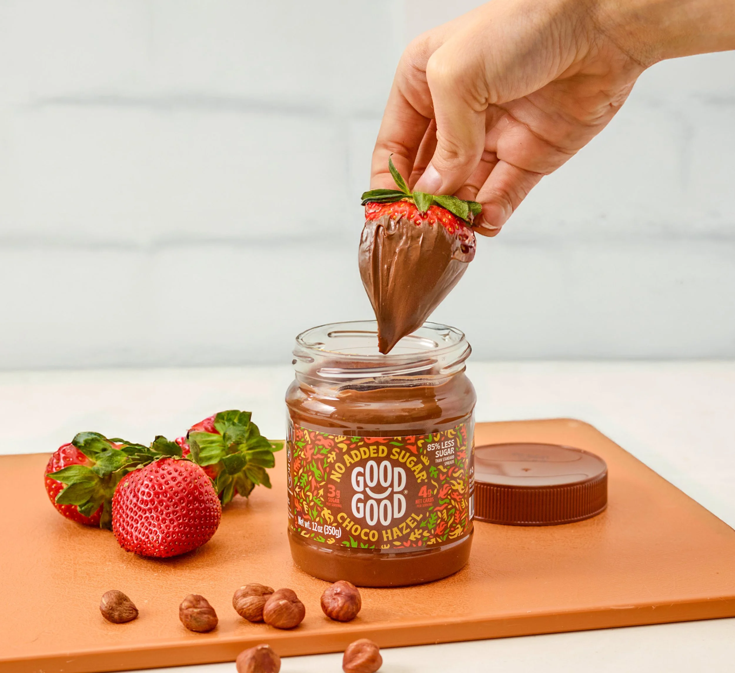 Hand dipping a strawberry into a jar of choco hazel spread with strawberries and hazelnuts on a cutting board.