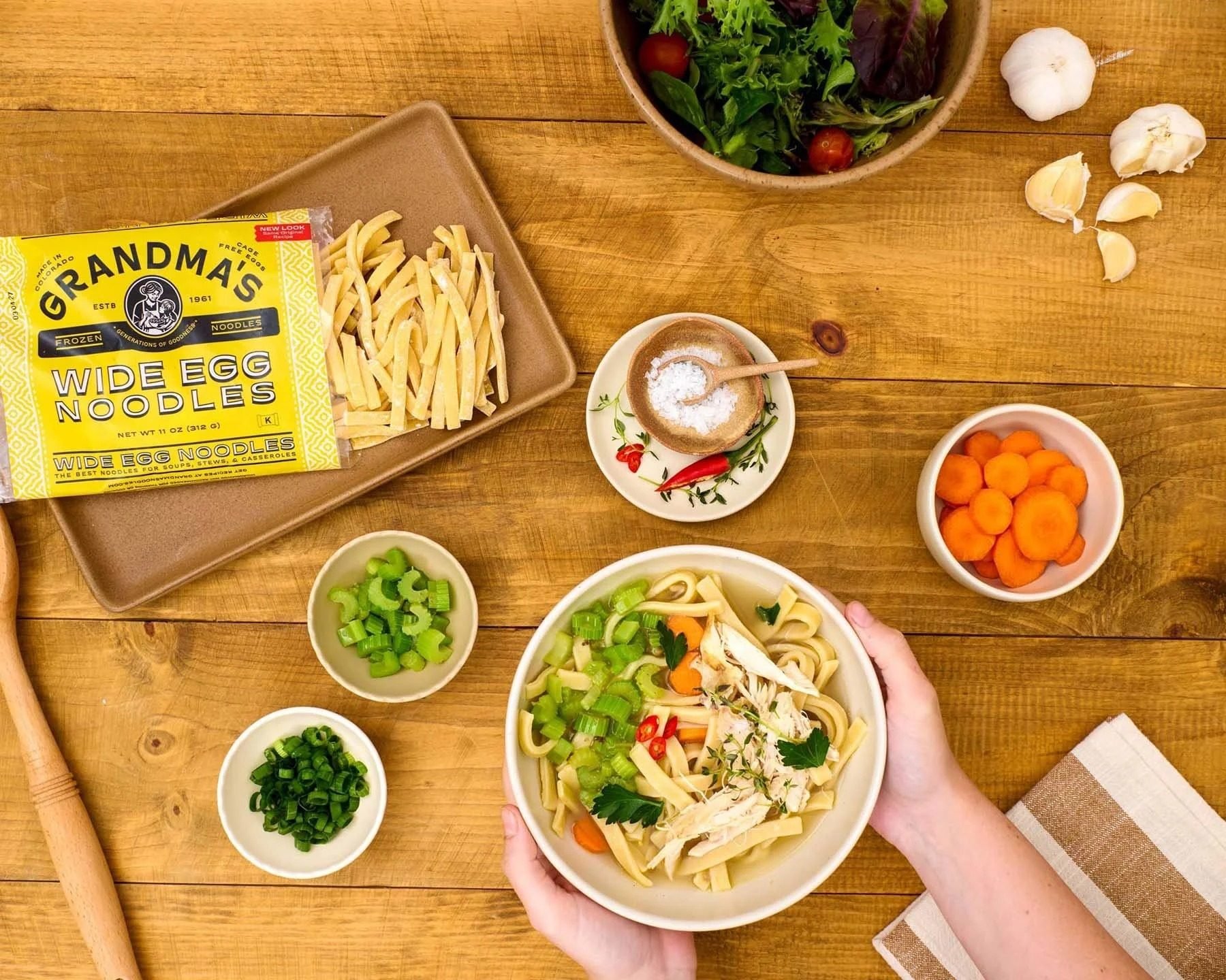 Grandma's noodles package with soup and hands on a wood table, CPG food photography