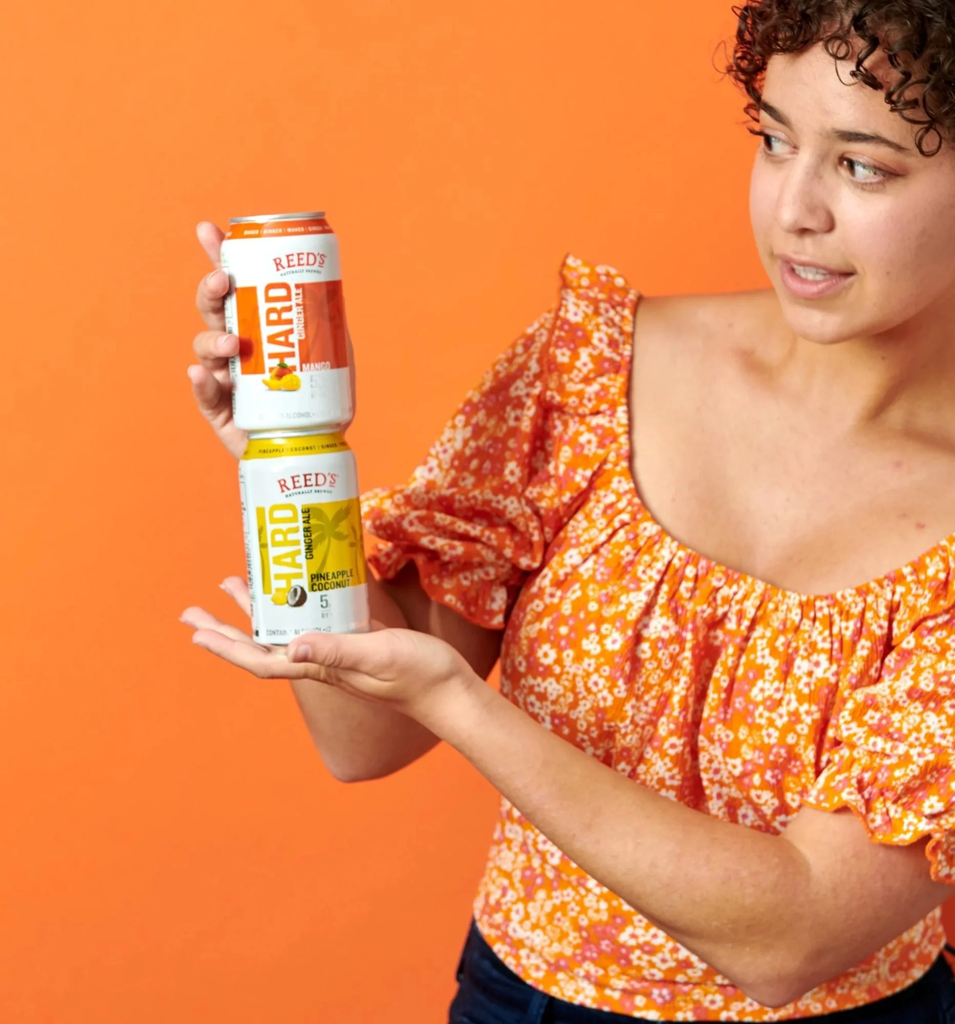 Model holding sparkling cans on orange in a floral top, CPG beverage lifestyle photography