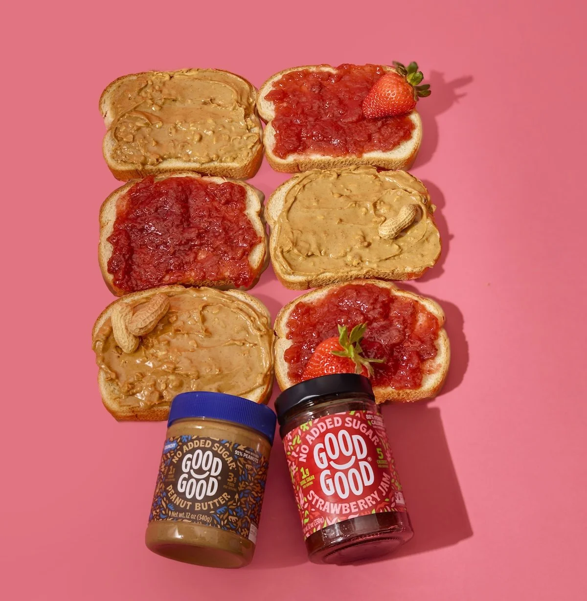 Six slices of bread topped with peanut butter and strawberry jam, two jars of peanut butter and strawberry jam, and two strawberries on a pink background.