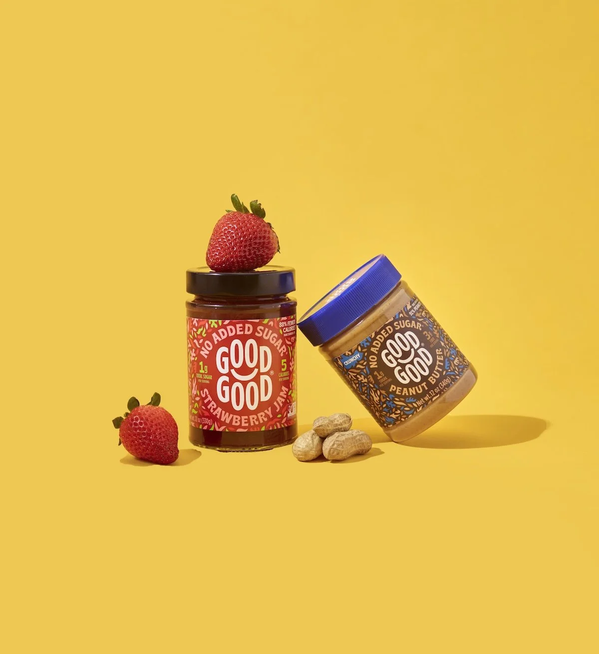 Two jars of Good Good brand nut butters and strawberry jam on a yellow background. One jar is upright with a strawberry on top, and the other jar is tilted. There are peanuts and a strawberry on the surface, casting shadows.