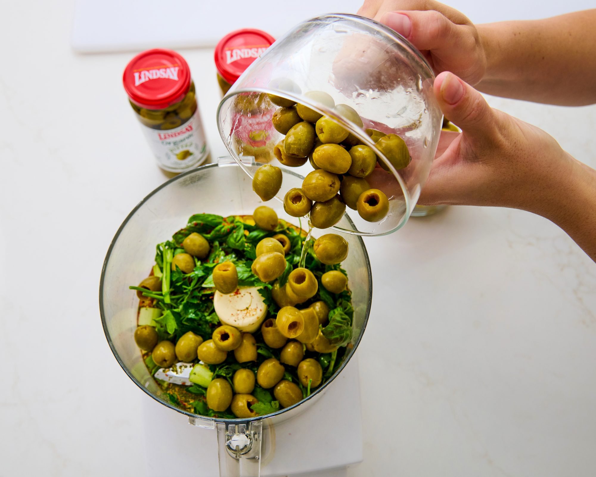 Green olives pouring into a food processor for a recipe step. Process photography for brand recipe content.