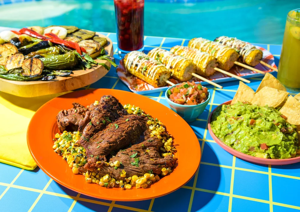 Grilled skirt steak and vegetables on an orange plate beside blue tile. Meat and grilling photography for summer campaigns.