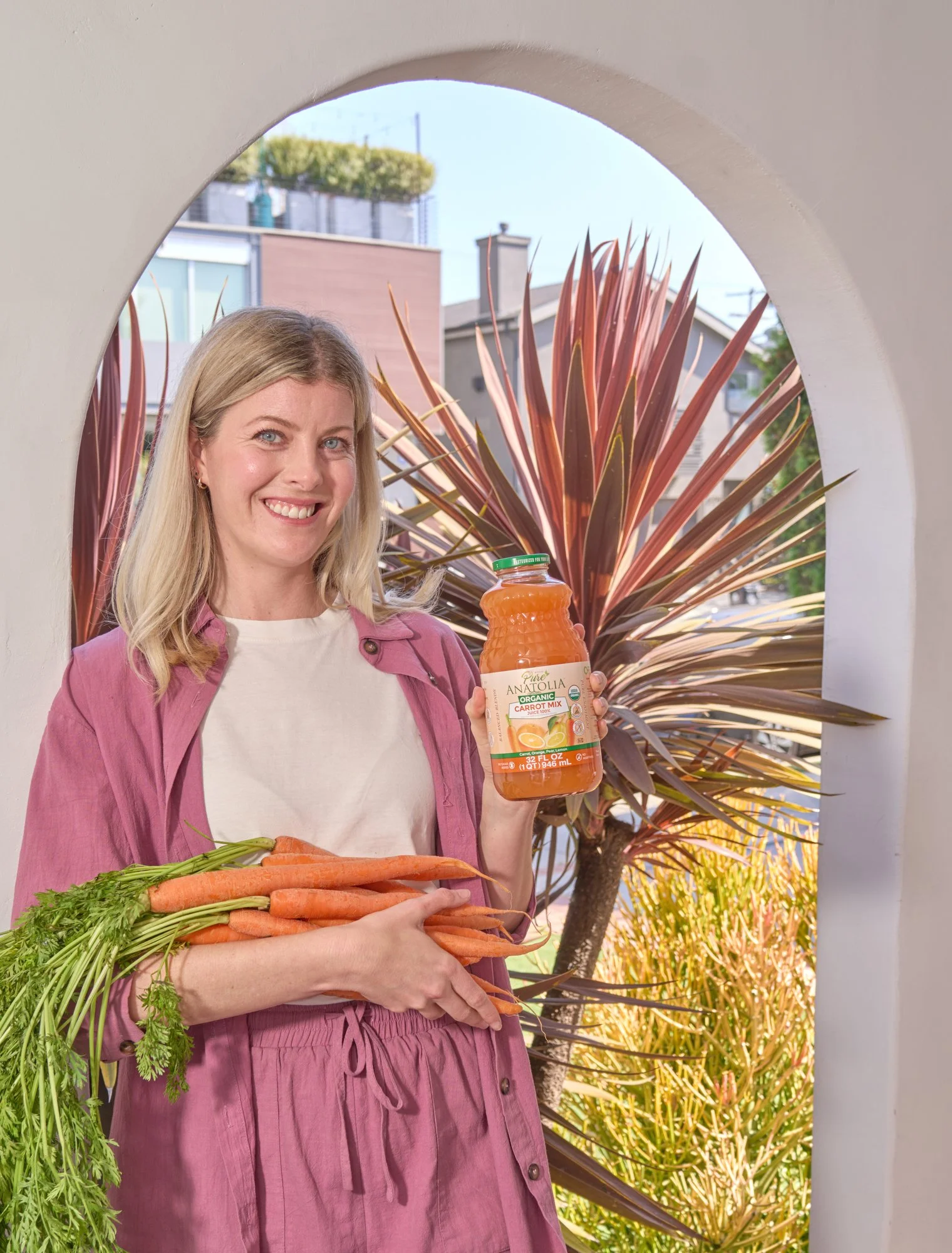 A woman smiling, holding fresh carrots in front of a decorative archway and large ornamental grass outdoors, with a background of buildings and sky.
