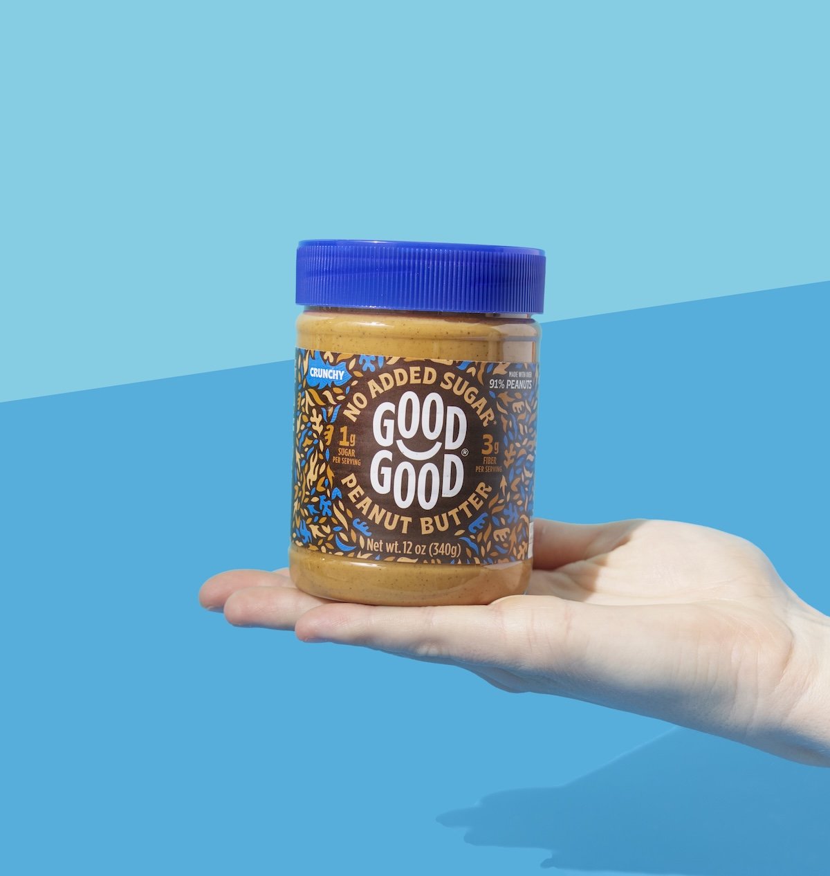 A hand holding a jar of Good Good peanut butter with a blue lid and a colorful label on a blue background.