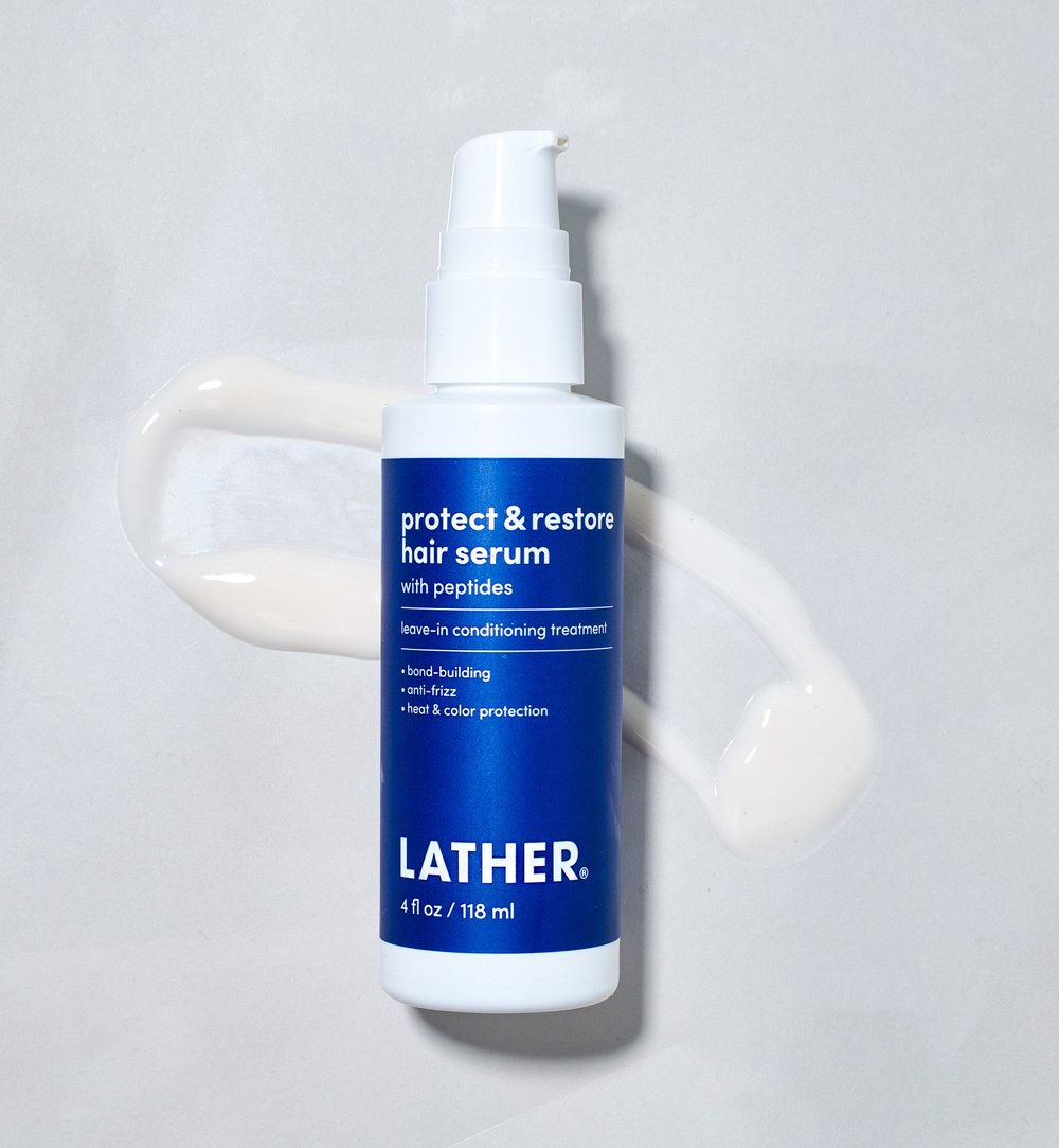 A white and blue bottle of LATHER Protect & Restore Hair Serum on a white surface with some serum spilled next to it.