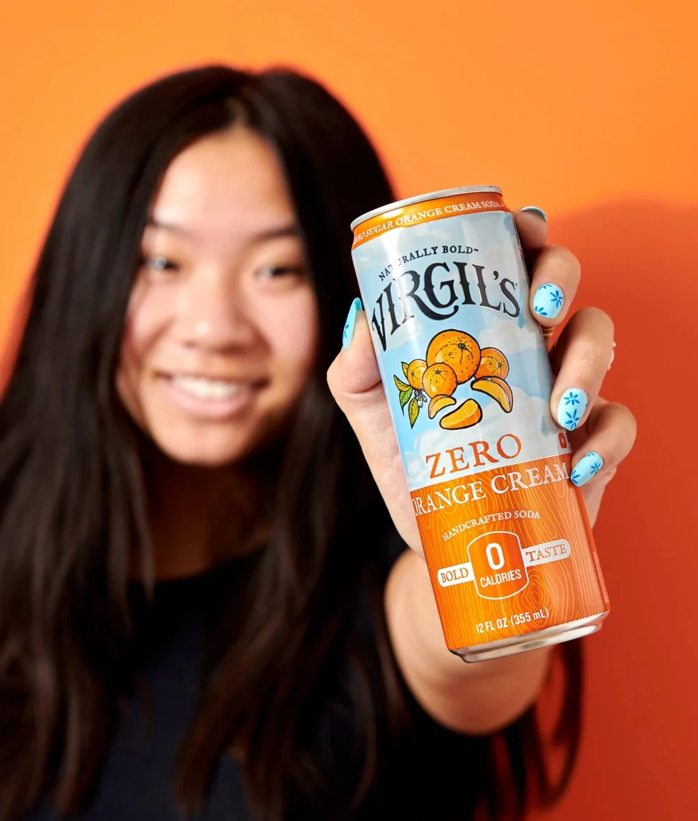 Smiling model holding a Virgil's Zero Sugar soda can toward camera on orange, CPG lifestyle photography