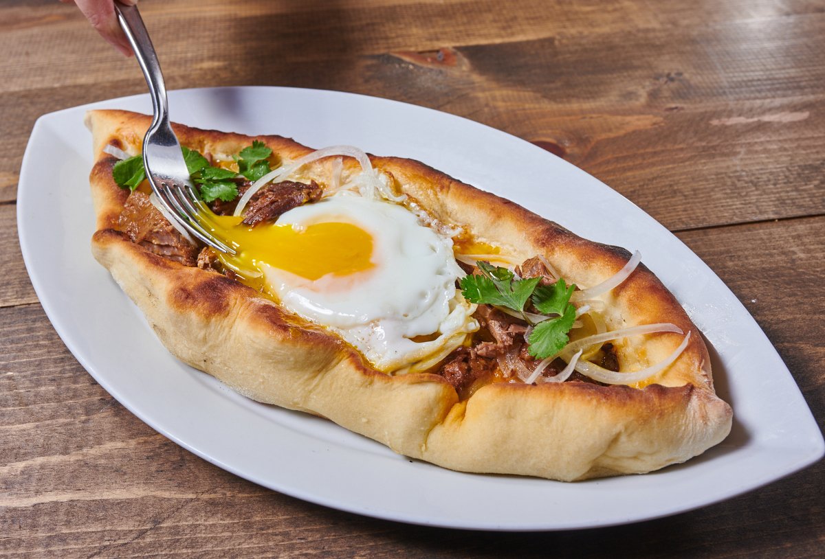 Khachapuri flatbread boat with a runny egg being cut, Culinary Institute of America restaurant menu
