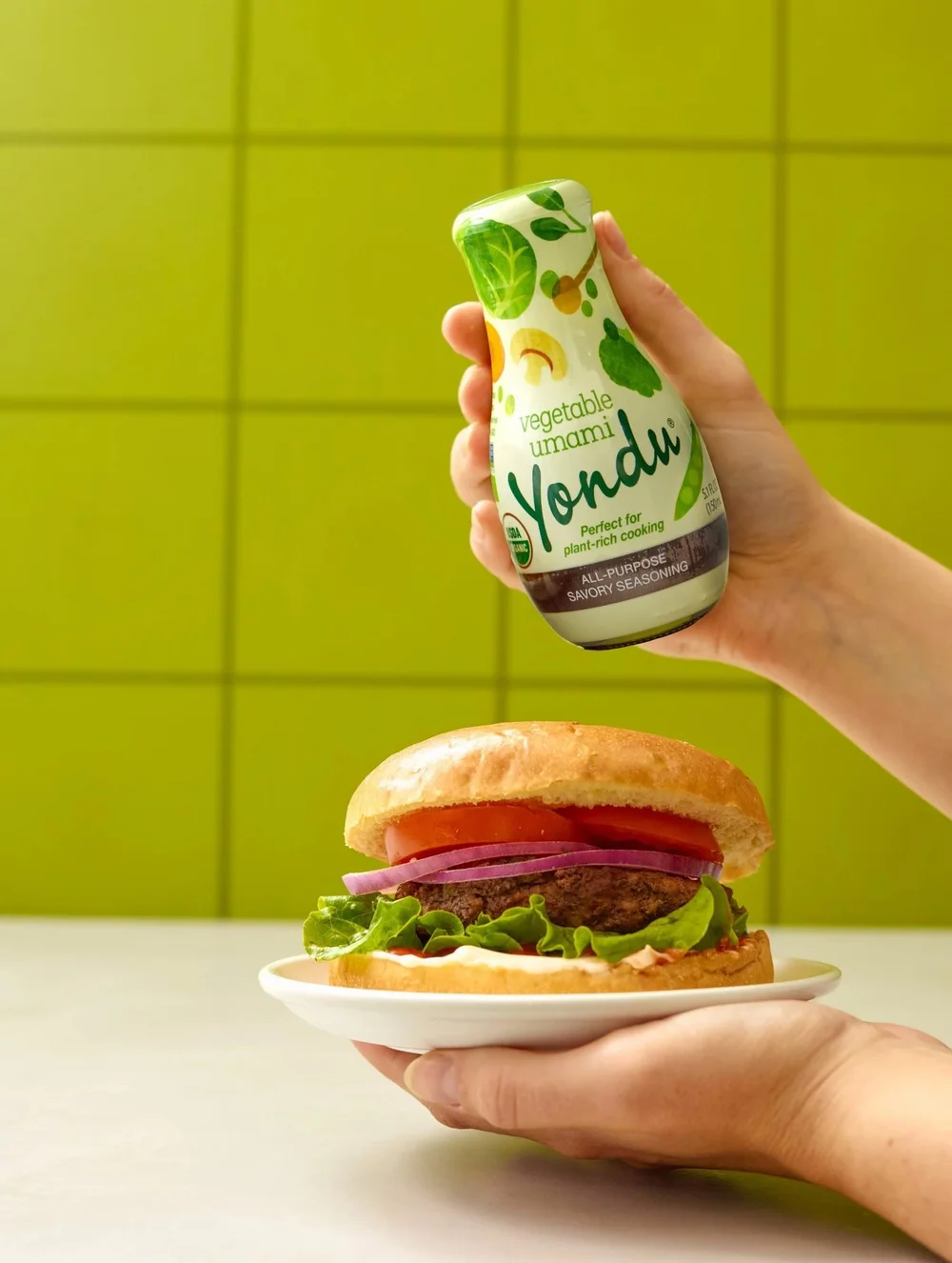 Hand holding a veggie burger beside a sauce bottle on green tile, lifestyle CPG food photography