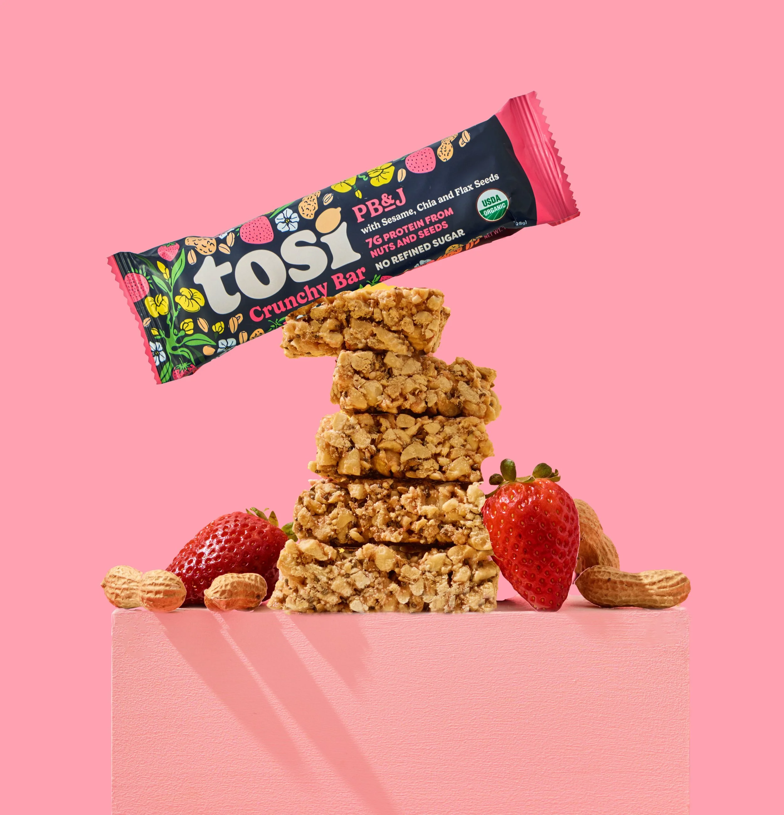 A stack of four granola bars with strawberries, peanuts, and almonds on a pink surface against a pink background, with a Tosi crunchy bar package floating above.