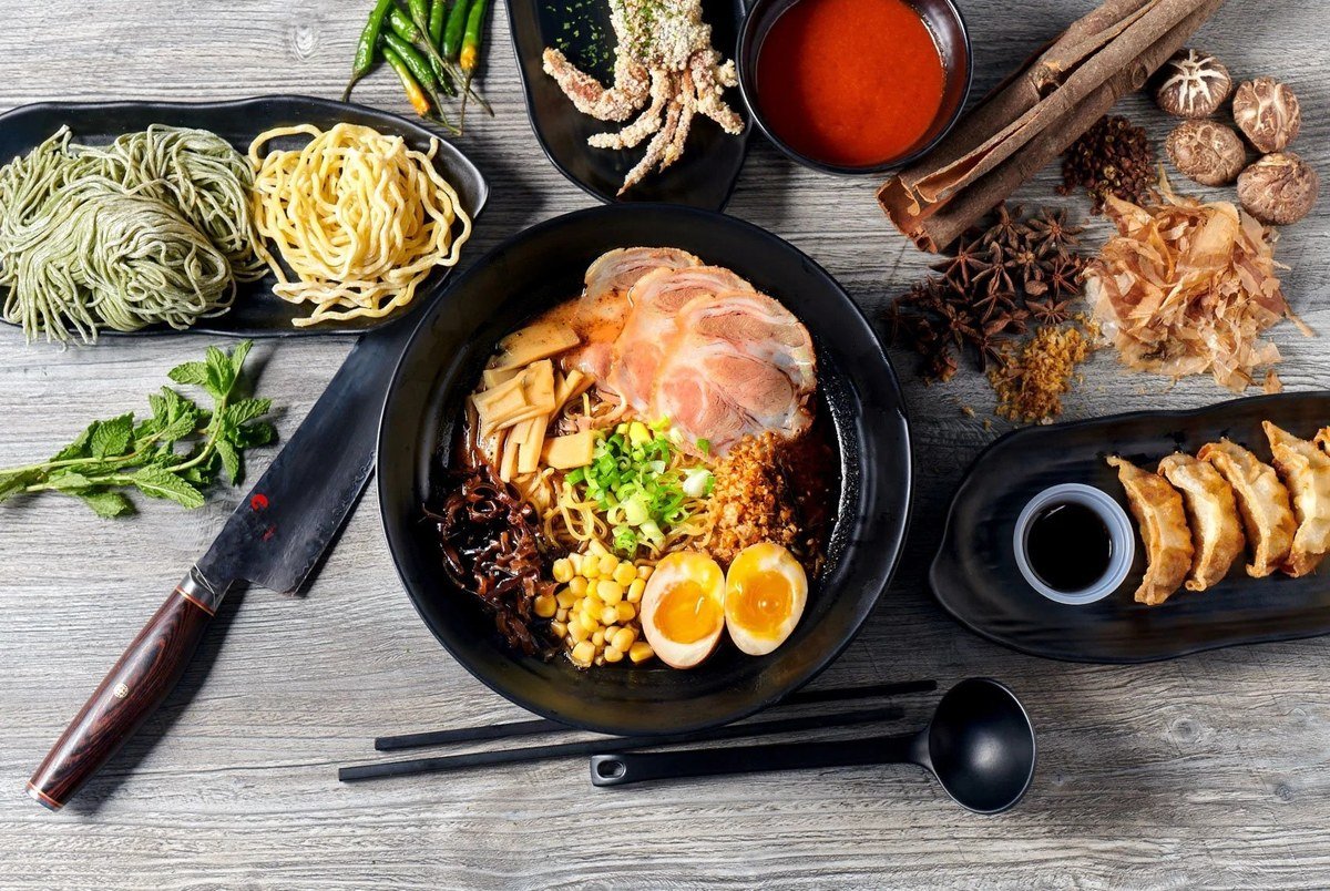 Ramen bowl with fresh ingredients and a chef's knife on a dark table