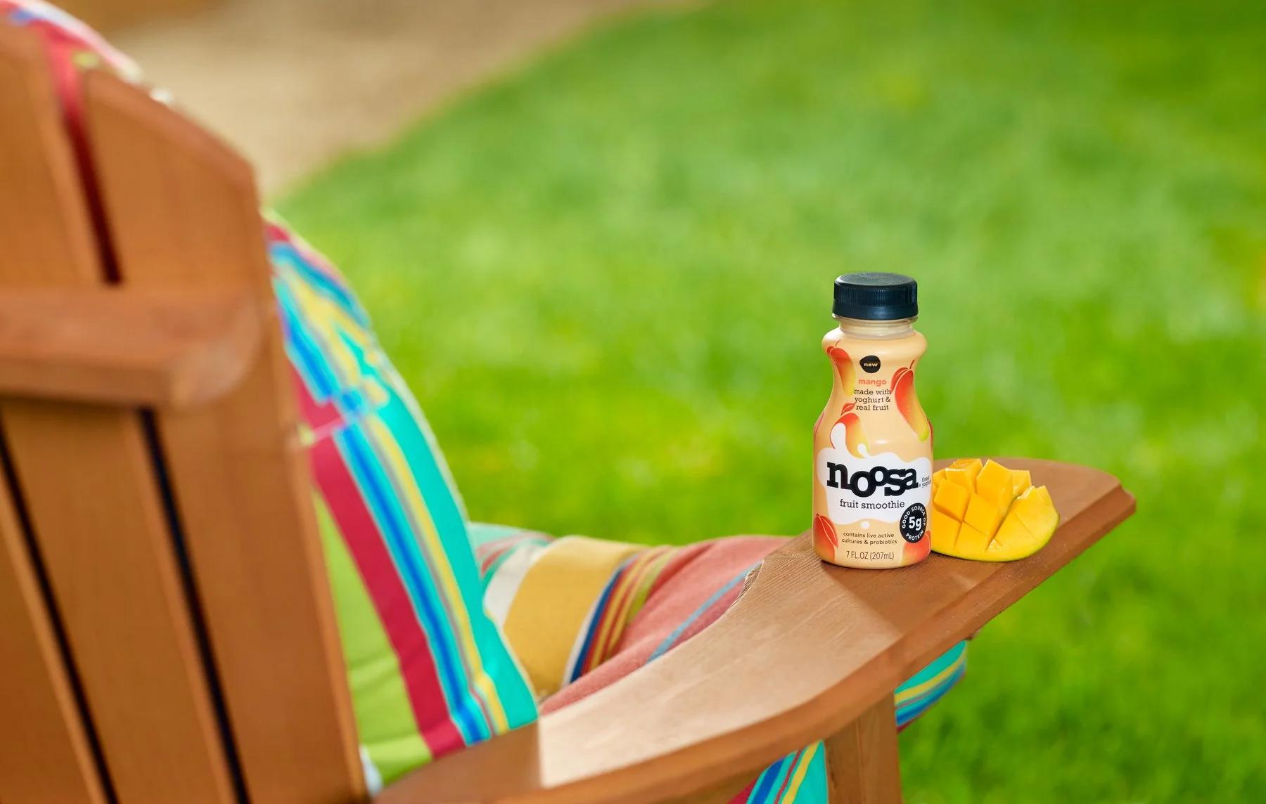A person sitting on a wooden outdoor chair with colorful striped fabric, with a bottle of Nosa fruit smoothie and sliced mango on a small wooden tray.