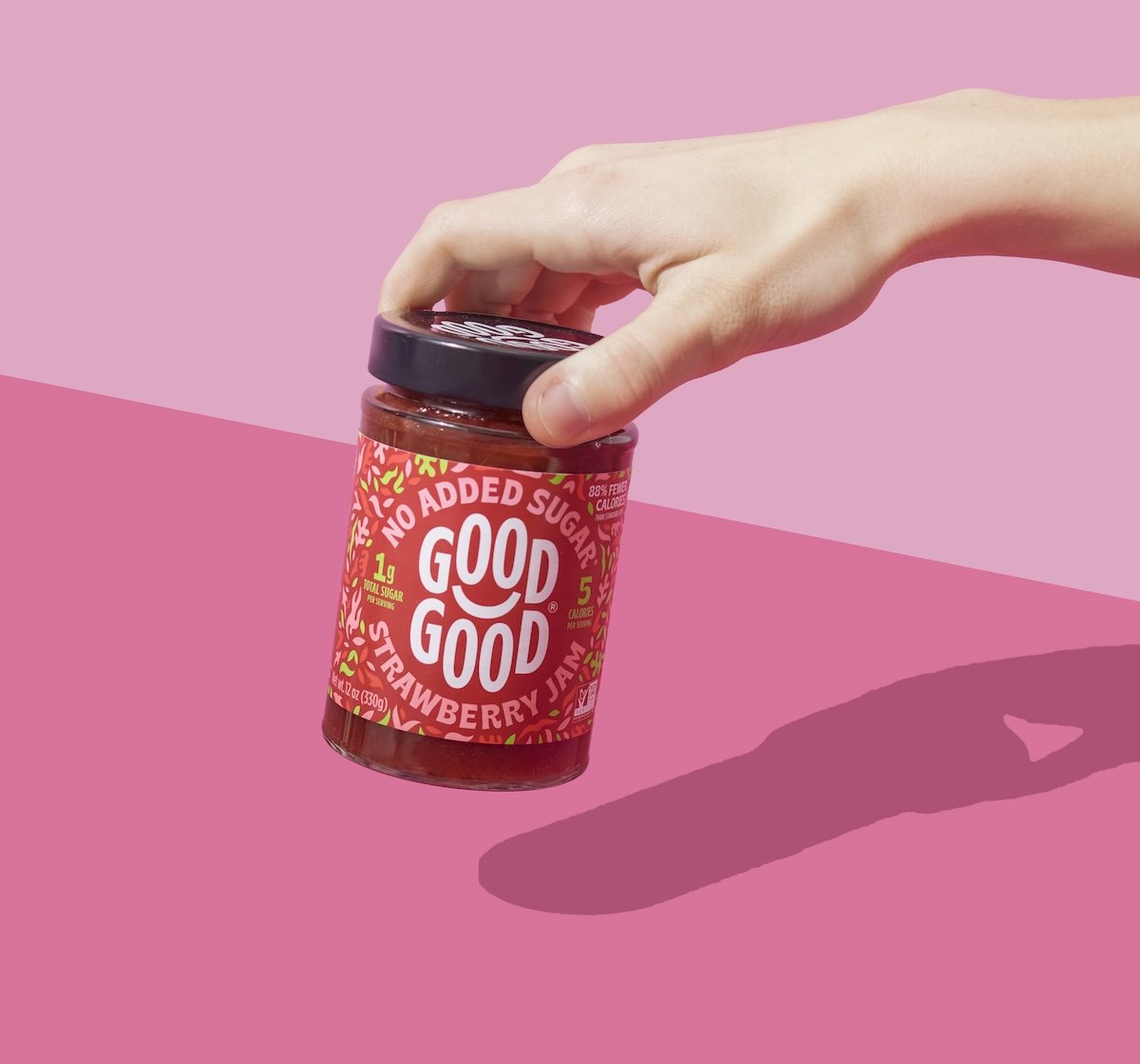 Hand holding a jar of Good Good strawberry jam against a pink background.