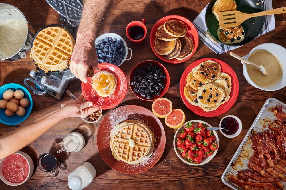 Overhead breakfast table of waffles, pancakes and fruit with hands. Overhead lifestyle food photography.