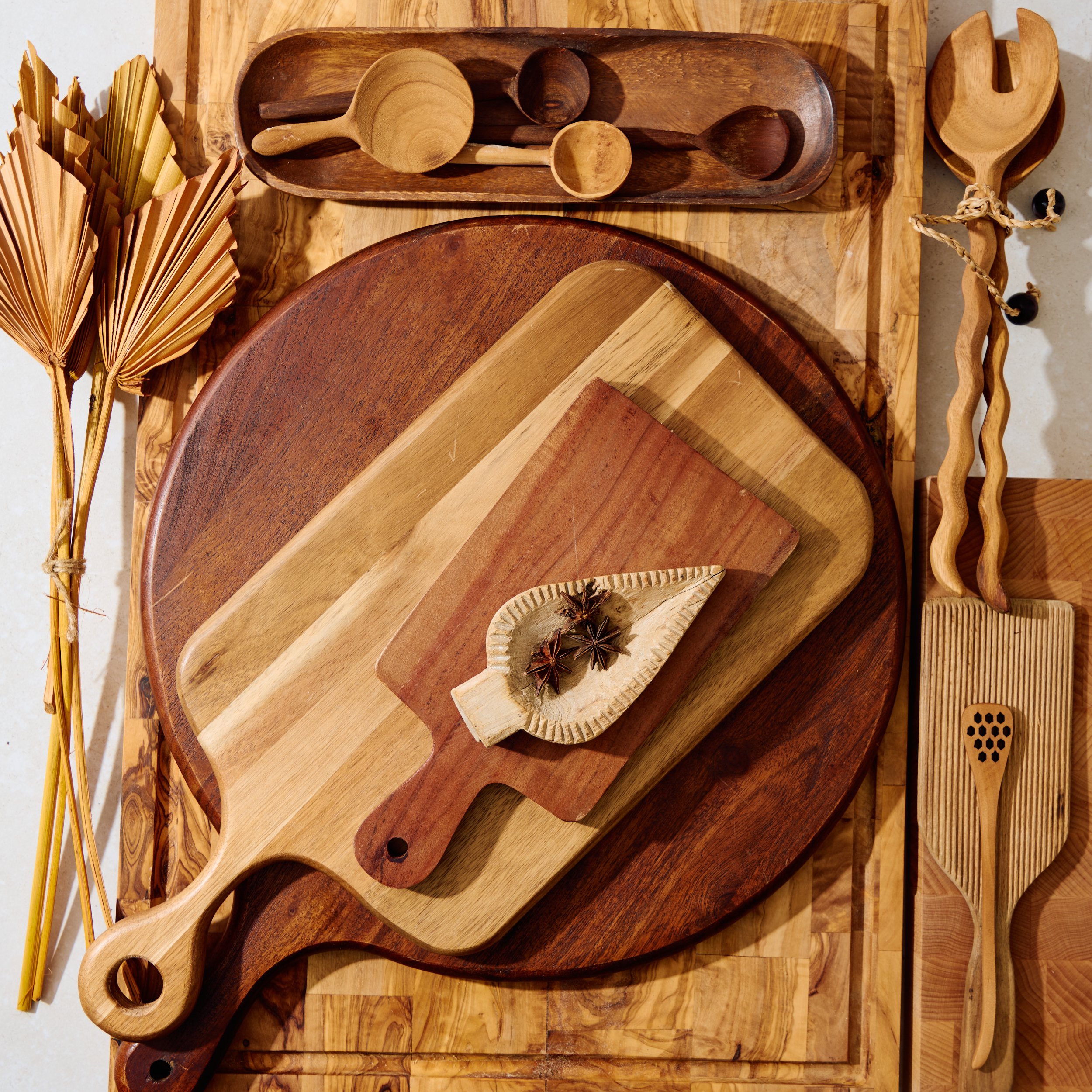 Arrangement of wooden cutting boards, utensils, and dried decorative plants on a wooden surface, featuring cutting boards of different sizes and shapes with dried star anise on top.