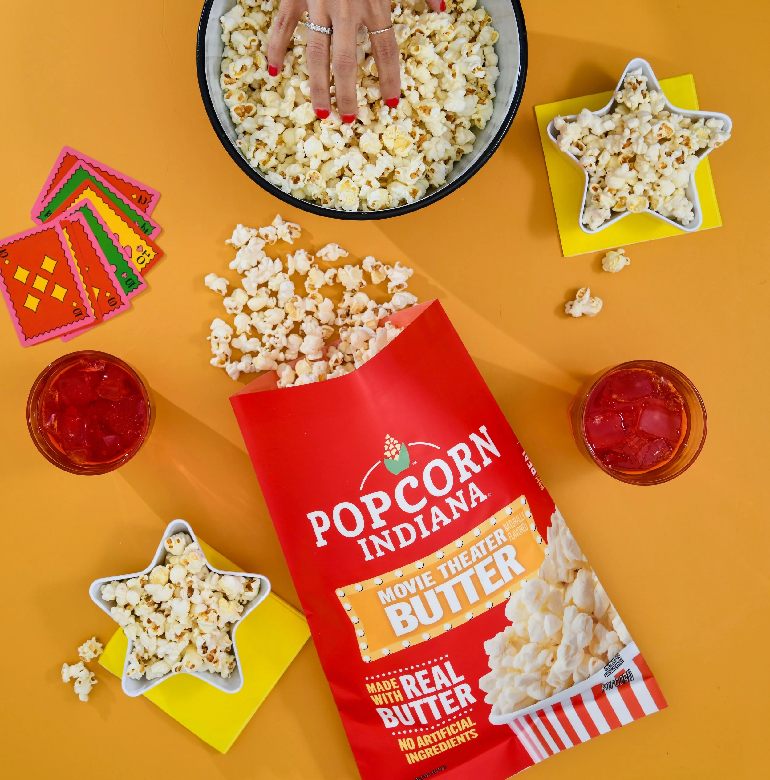 Popcorn in a red and white bag with yellow and white signs reading "Movie Theater Flavored Butter" and "Made with Real Butter," two glasses of iced red beverages, star-shaped bowls of popcorn, and colorful scratch-off tickets on an orange table.