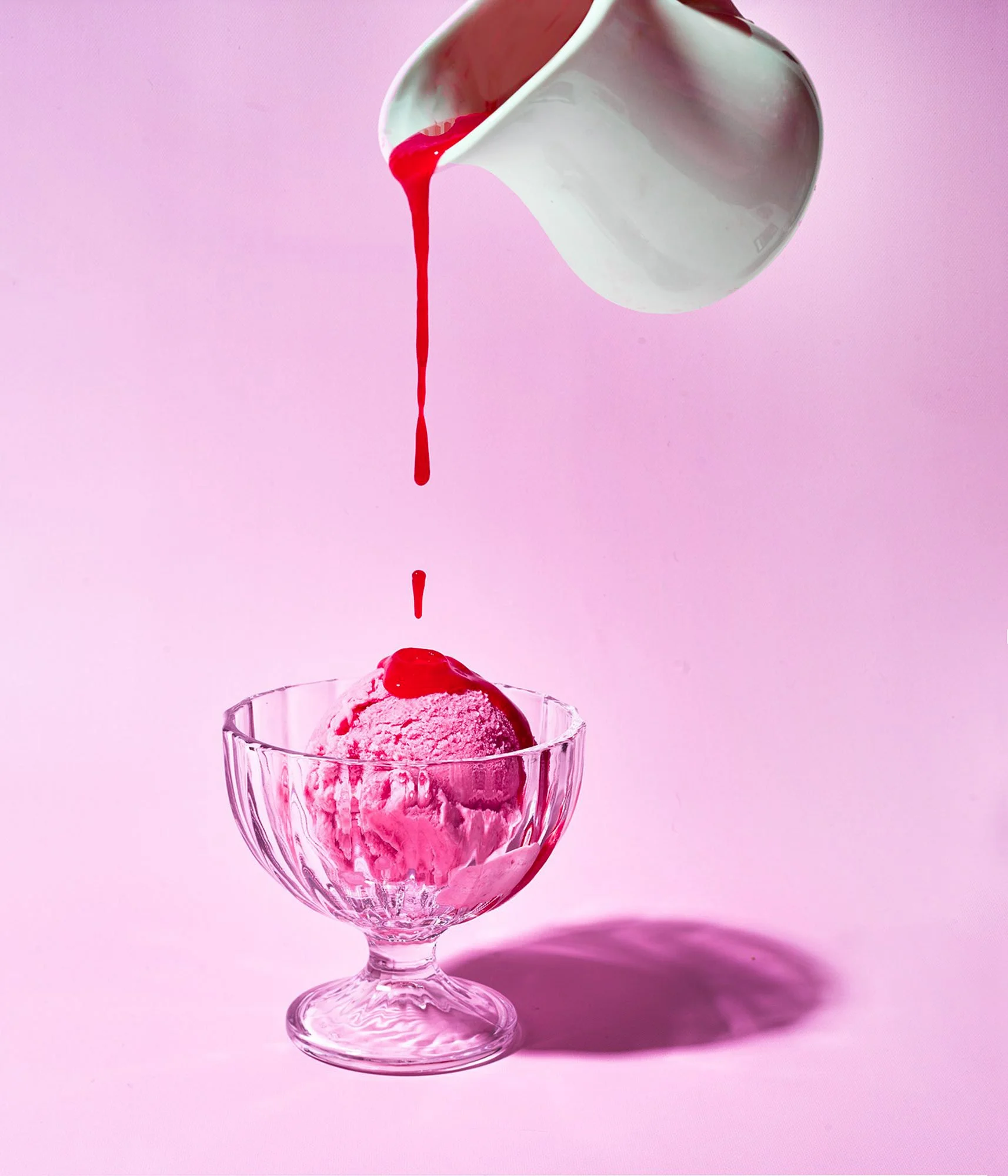Raspberry sauce pouring over ice cream in a glass coupe on pink. Action food photography for dessert and topping brands.