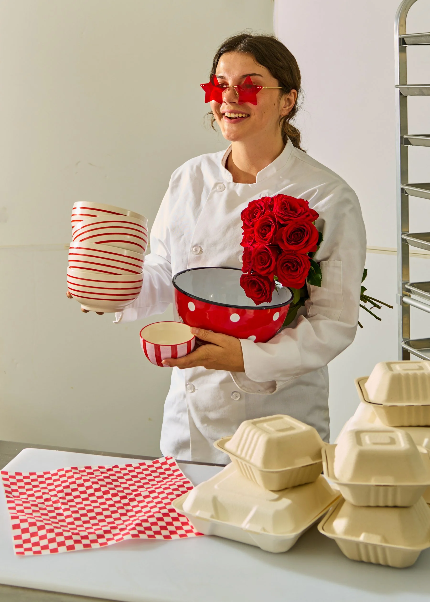A woman in a chef's coat wearing star-shaped red sunglasses, holding a bouquet of red roses and red-and-white polka dot bowl, standing behind a table with takeout boxes, in a commercial kitchen setting.