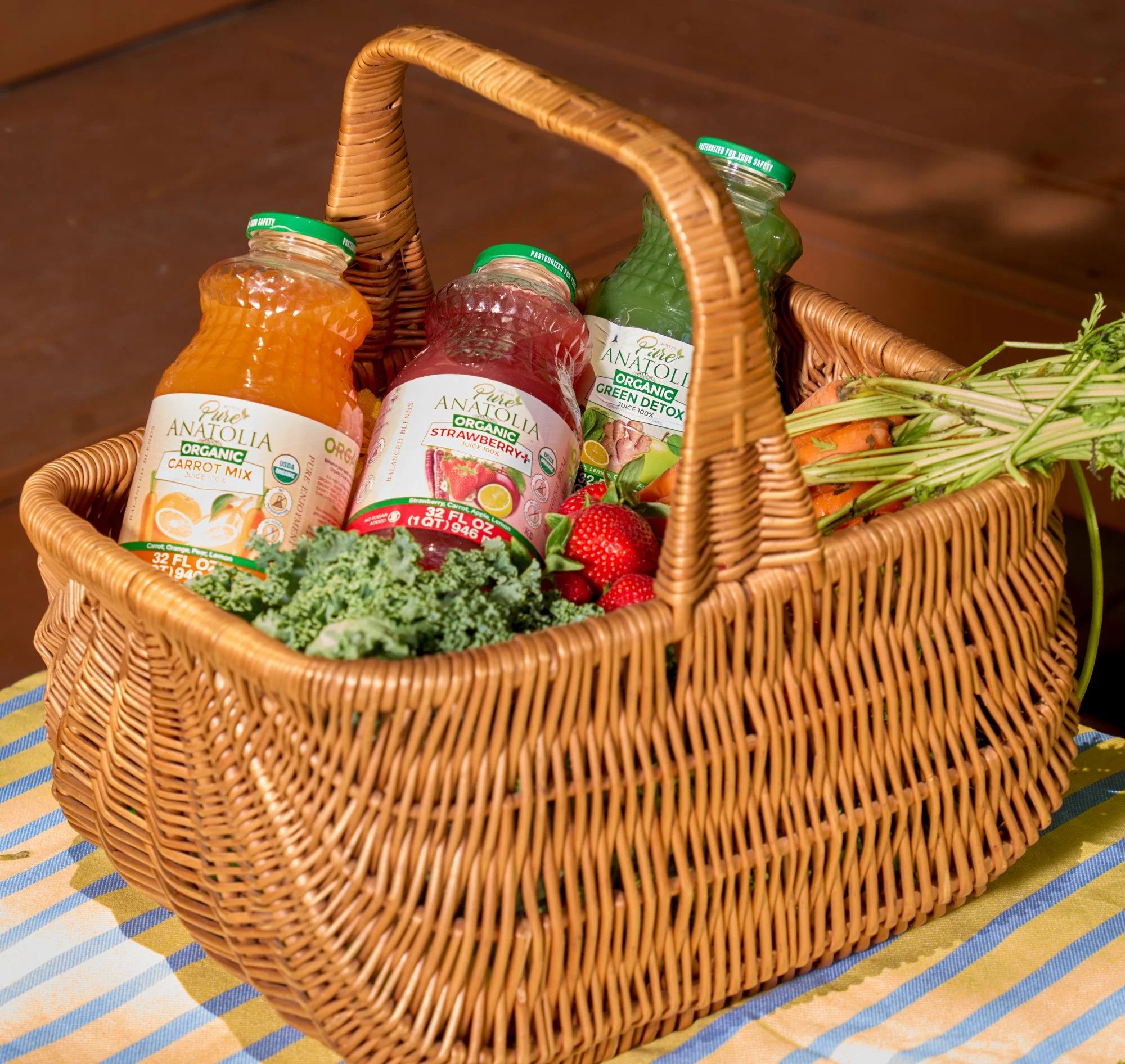 Wicker basket filled with jars of organic juice, including carrot mix, strawberry, and green detox, placed on a pastel striped tablecloth.