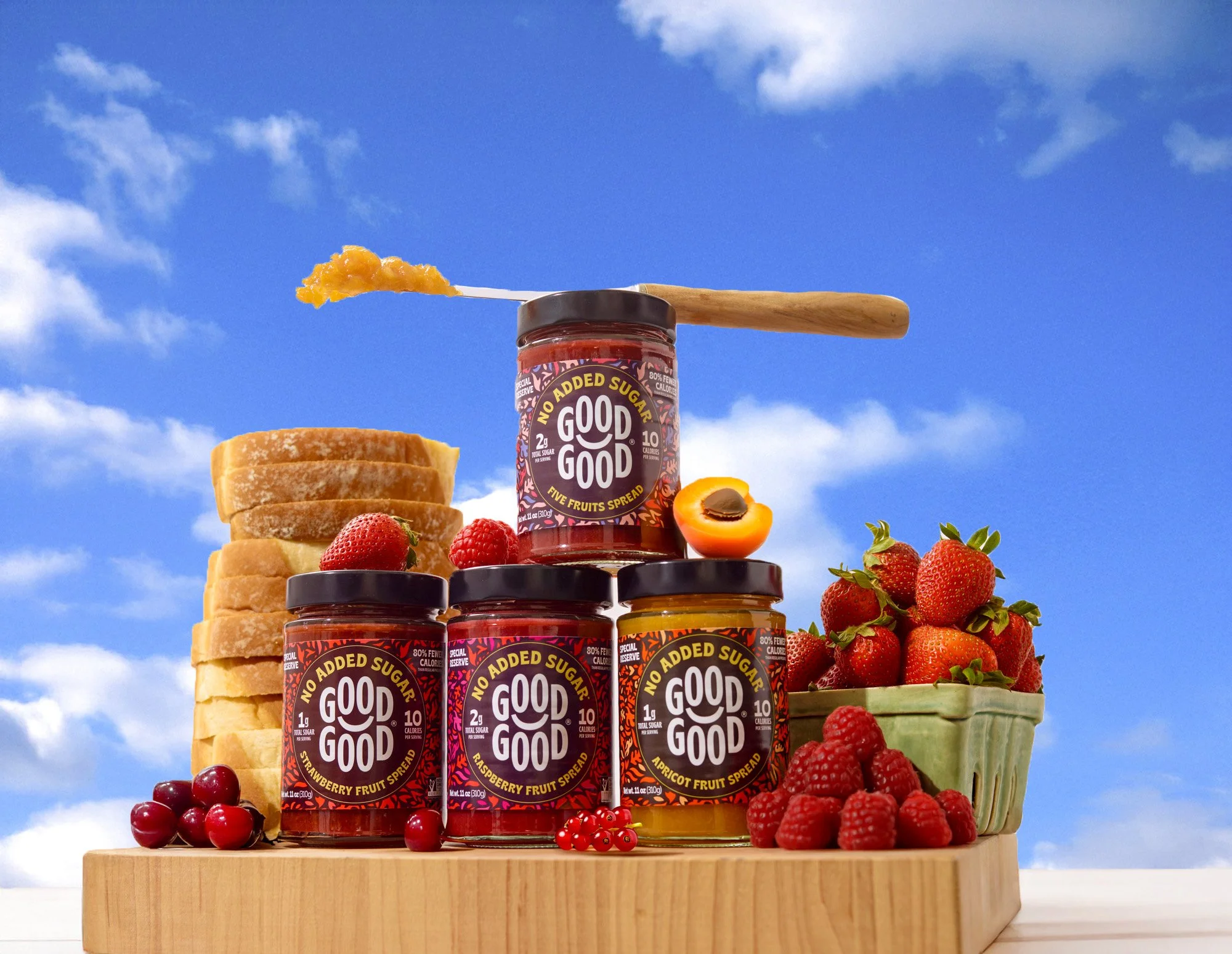 Stacks of sliced bread, jars of fruit preserves labeled 'Good Good' with no added sugar, fresh strawberries, raspberries, cherries, an apricot half, and a bowl of strawberries arranged on a wooden surface against a sky background.